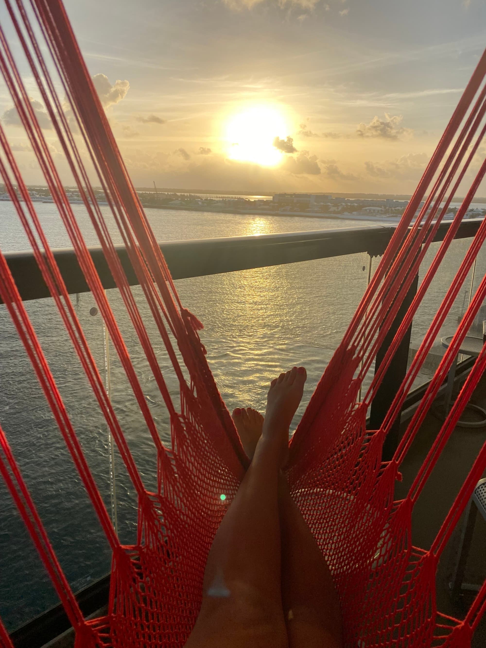 A picture of a persons legs outstretched on a red hammock, lounging beneath the bright sun. There is water in the distance.