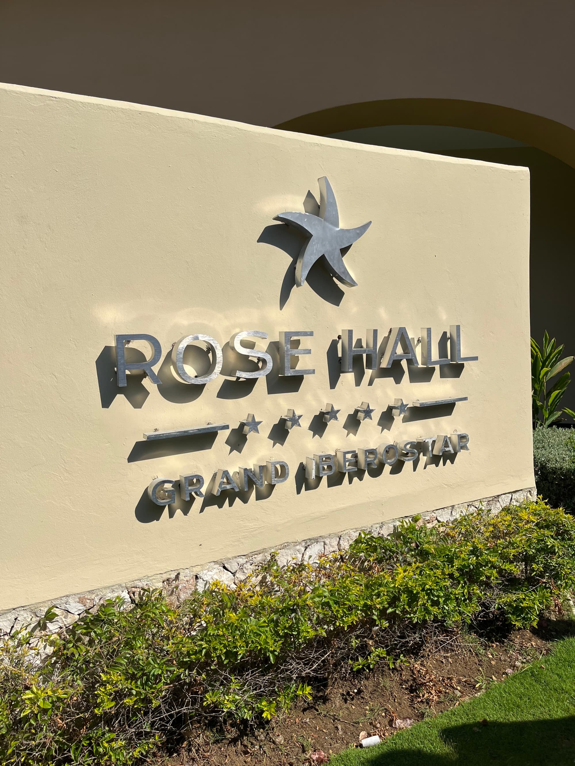 A sign that reads "Rose Hall Grand Iberostar" in chrome lettering above a set of bushes.