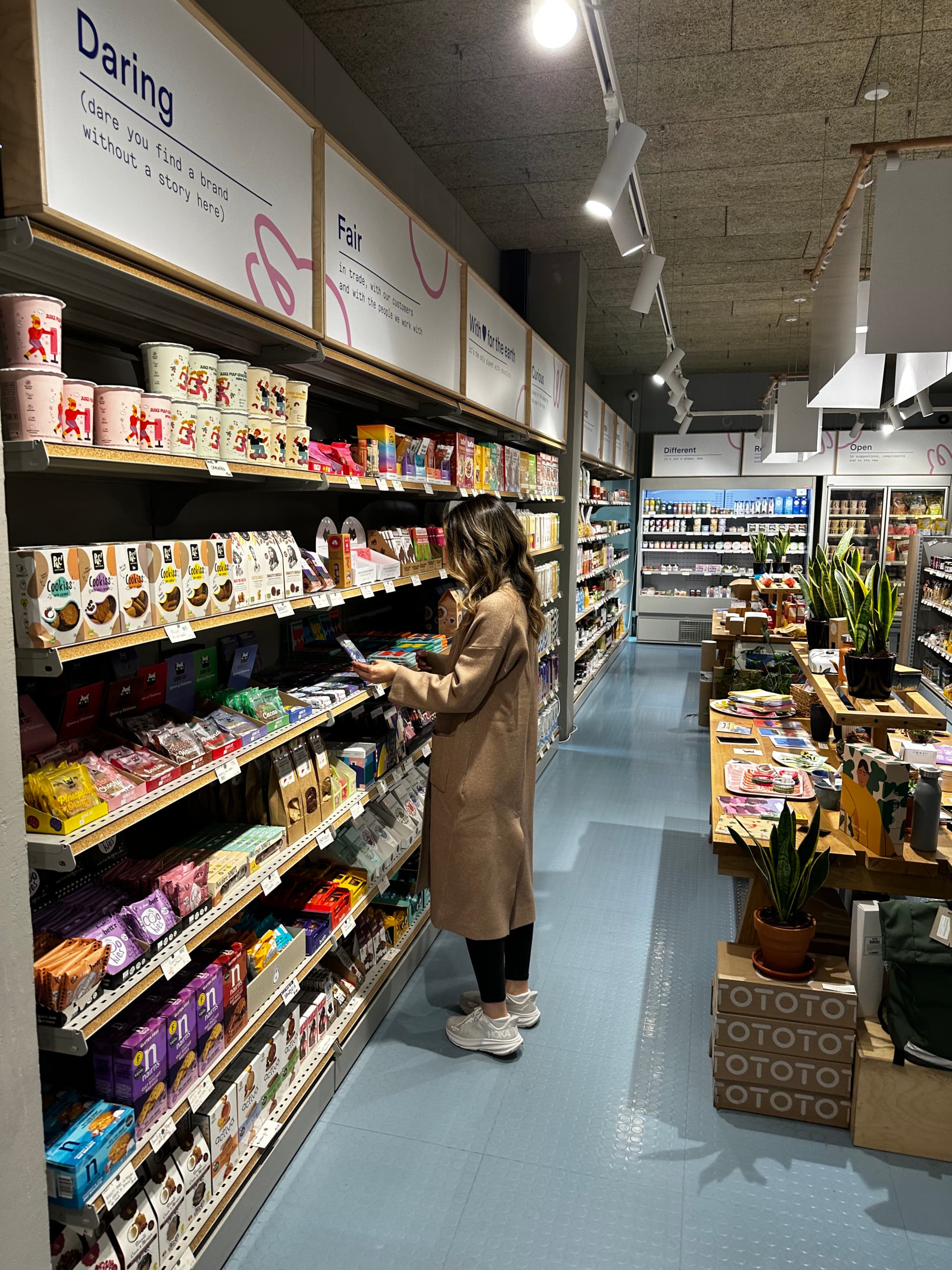 A person wearing a long coat and tennis shoes, holding a piece of merchandise inside of a supermarket.