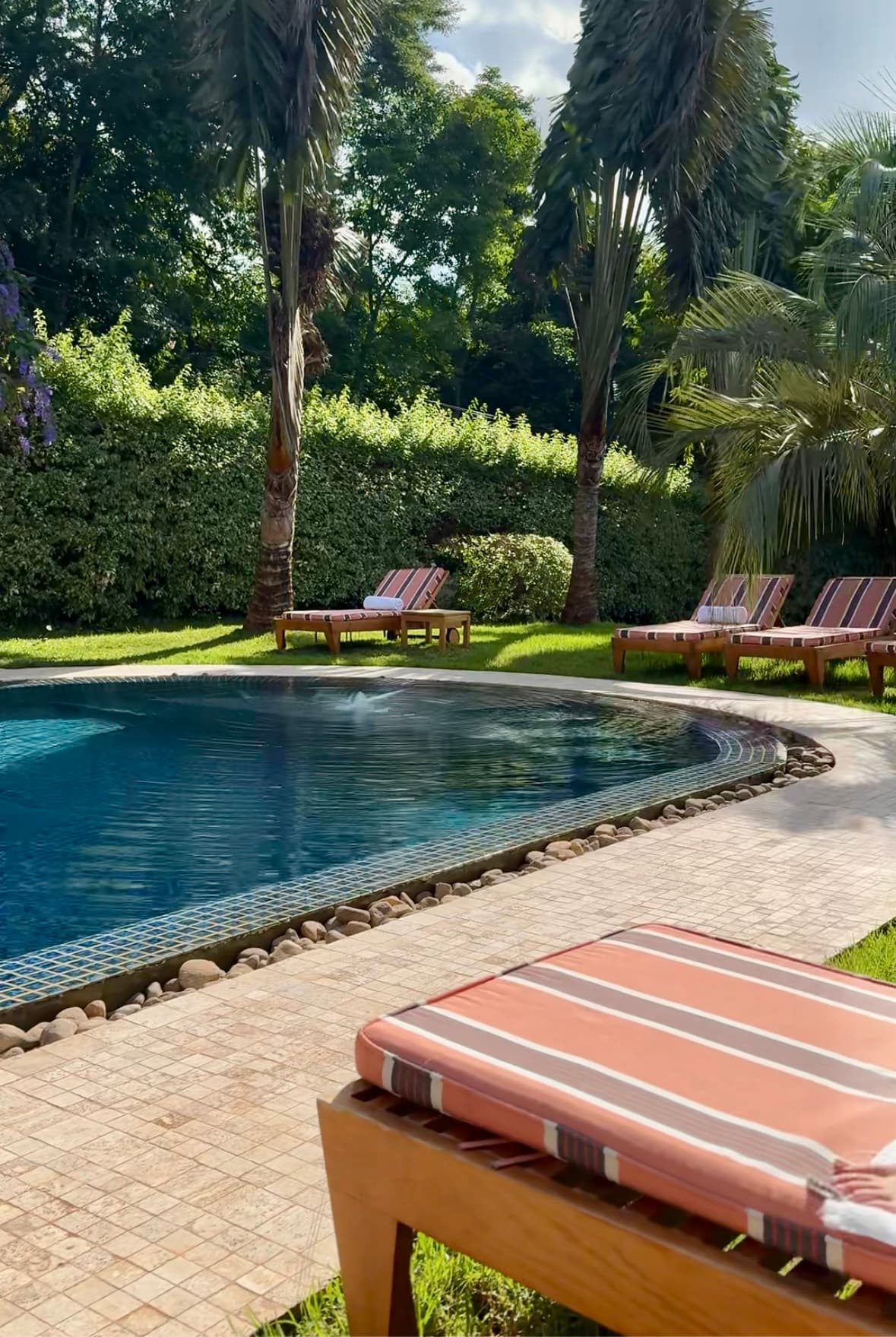 A lounge chair with a striped cushion in front of a swimming pool surrounded by more lounge chairs, grass and trees.