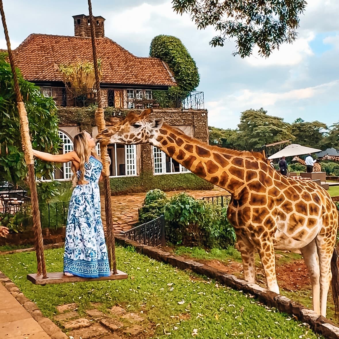 A woman posing in a blue and white patterned dress, next to large giraffe. There is a brown house with white paned windows and trees in the background.