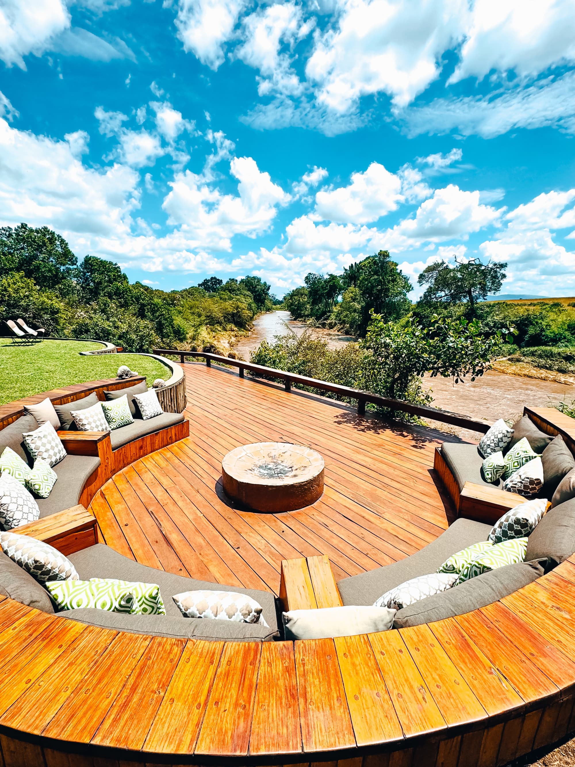 An wooden patio with couches, a fire pit, trees and a cloudy blue sky overhead.