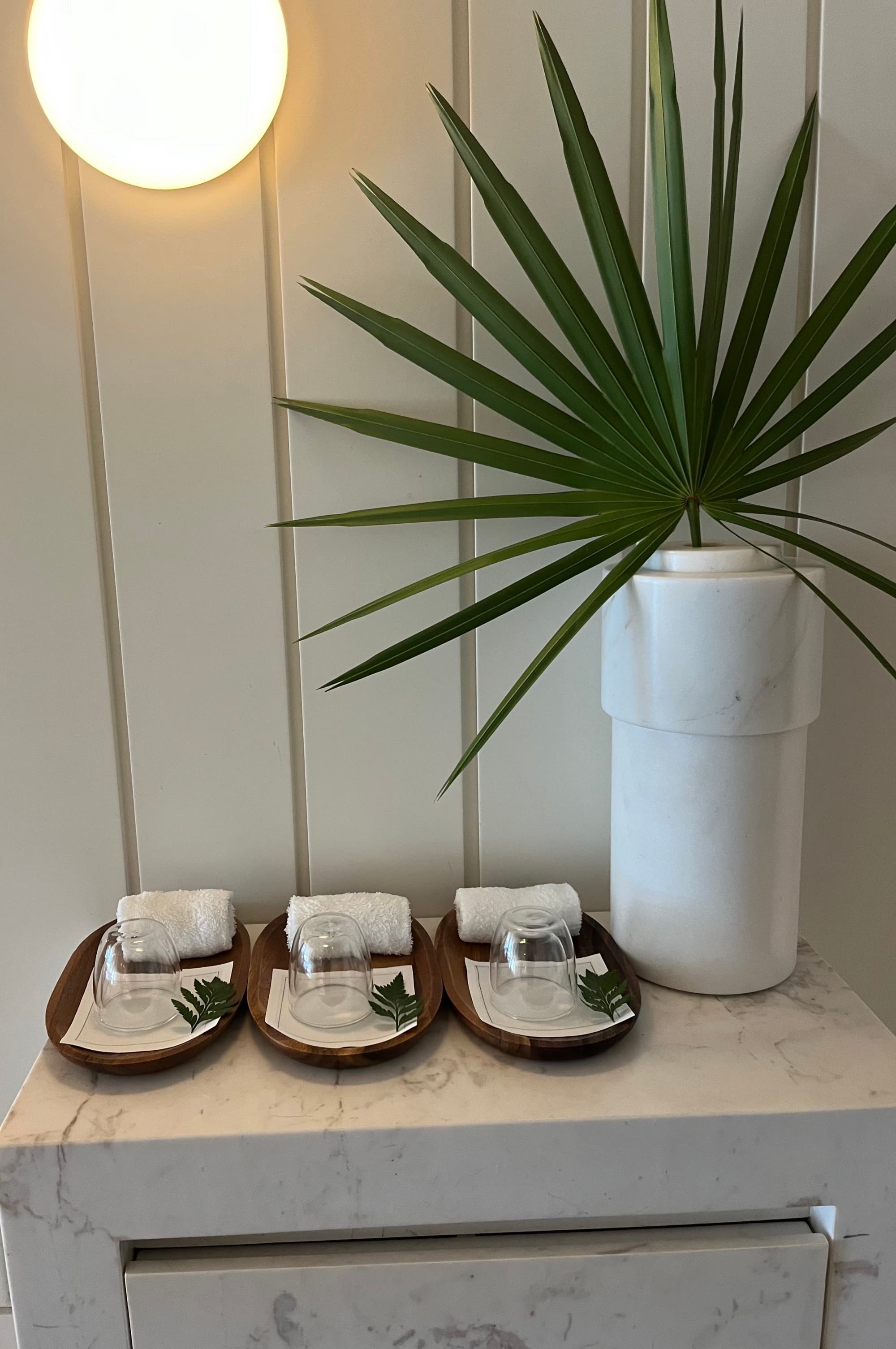 A palm leaf in a white vase placed on top of a marble table with white paneled walls behind it. There is a round sconce turned on and treats next to the vase.