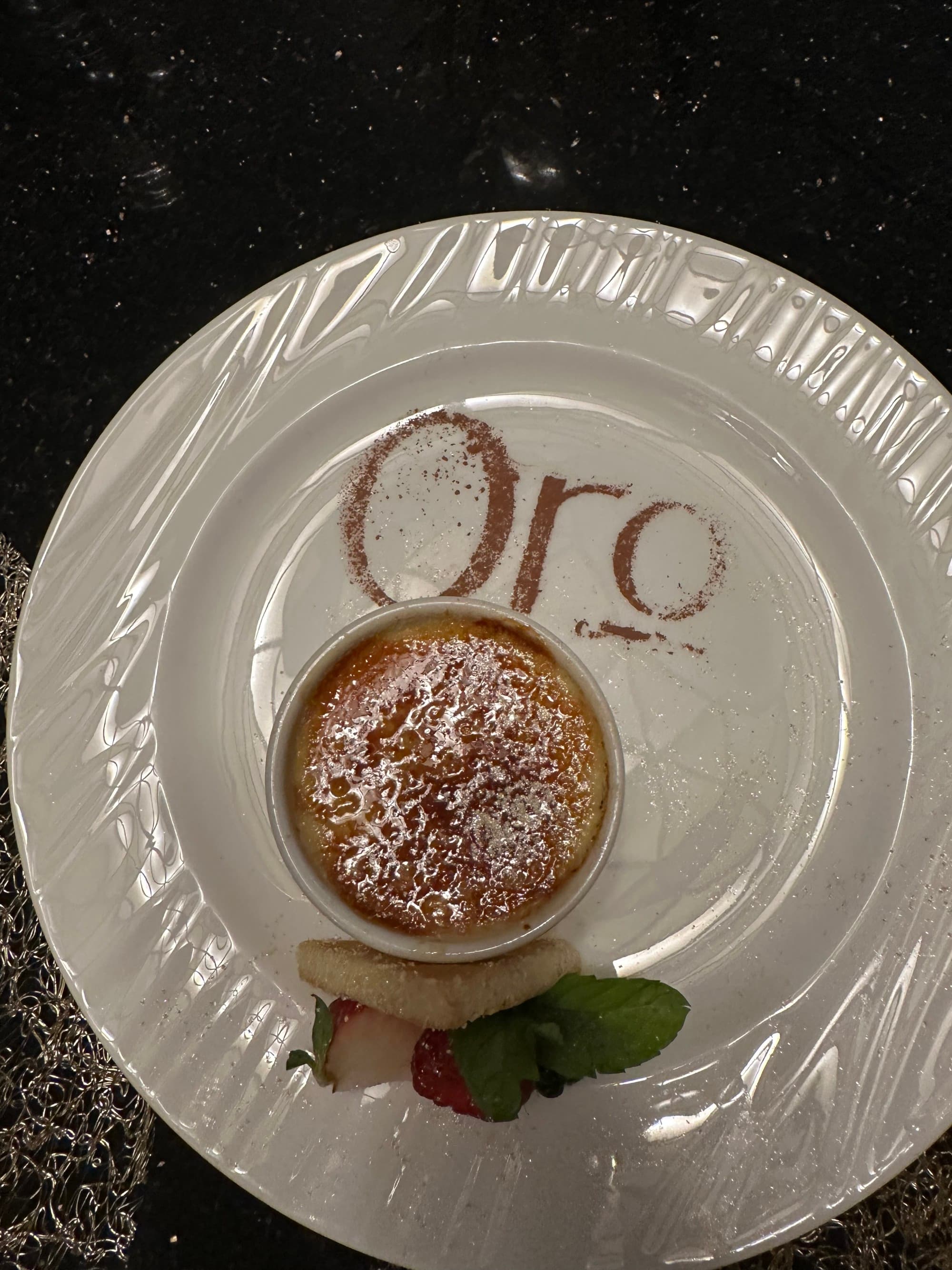 A white plate with crème brûlée and fruit on top of it next to edible letters that read "Oro" on the top of the plate.