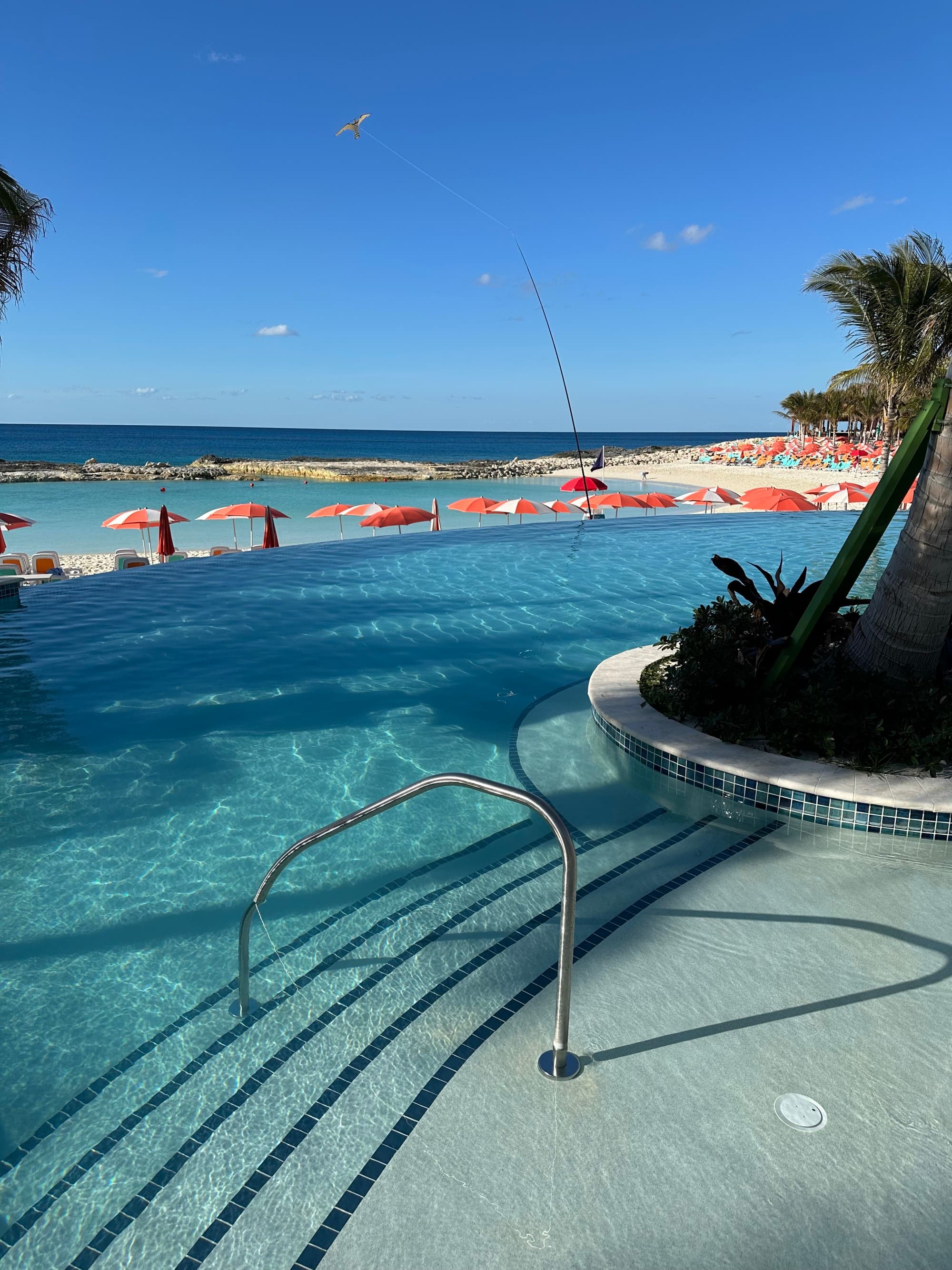 A pool behind the ocean with orange umbrellas in between.