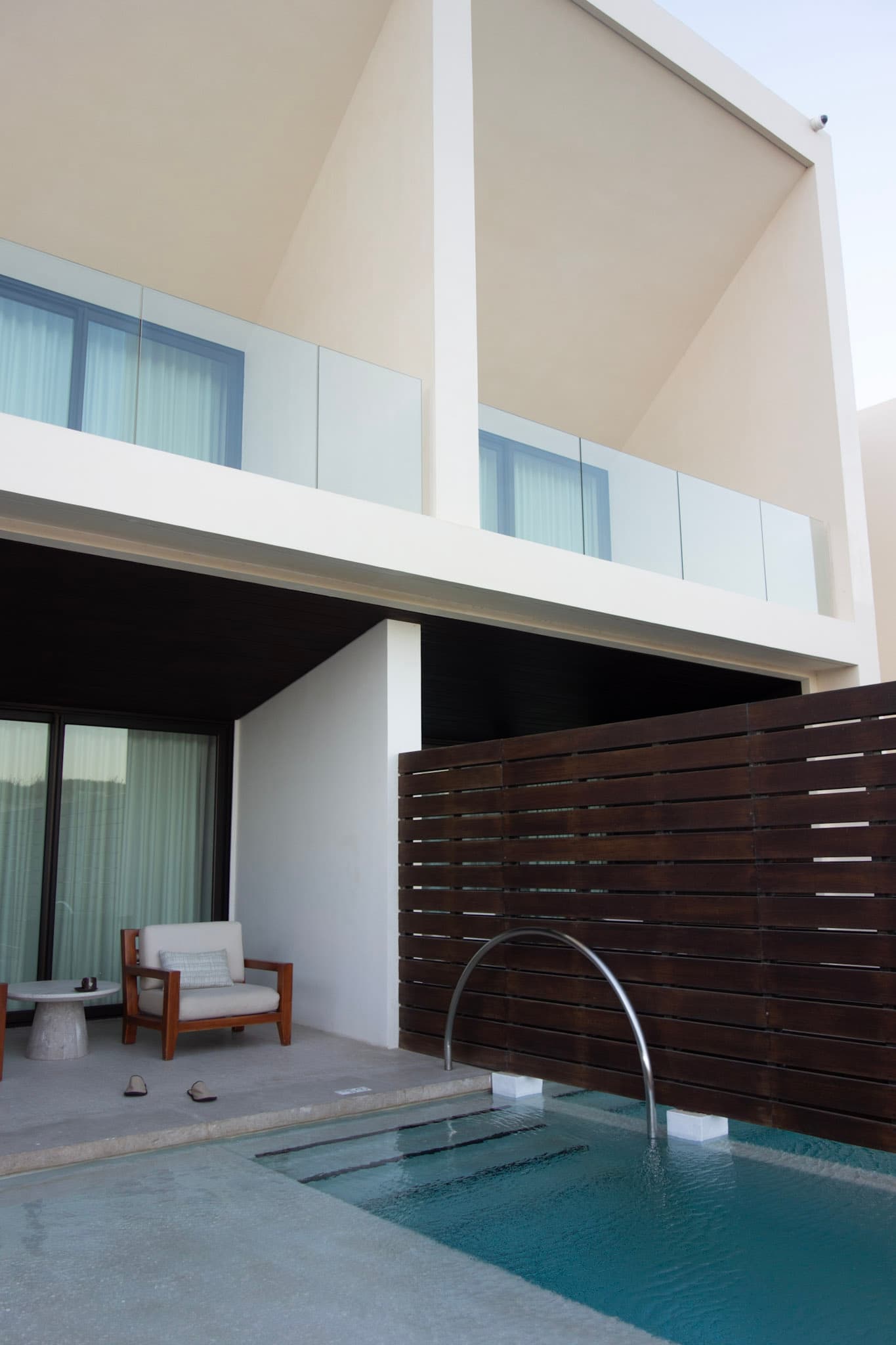 A view of a hotel suite pool with a white lounge chair, wooden privacy wall and glass walled balconies on the second level.