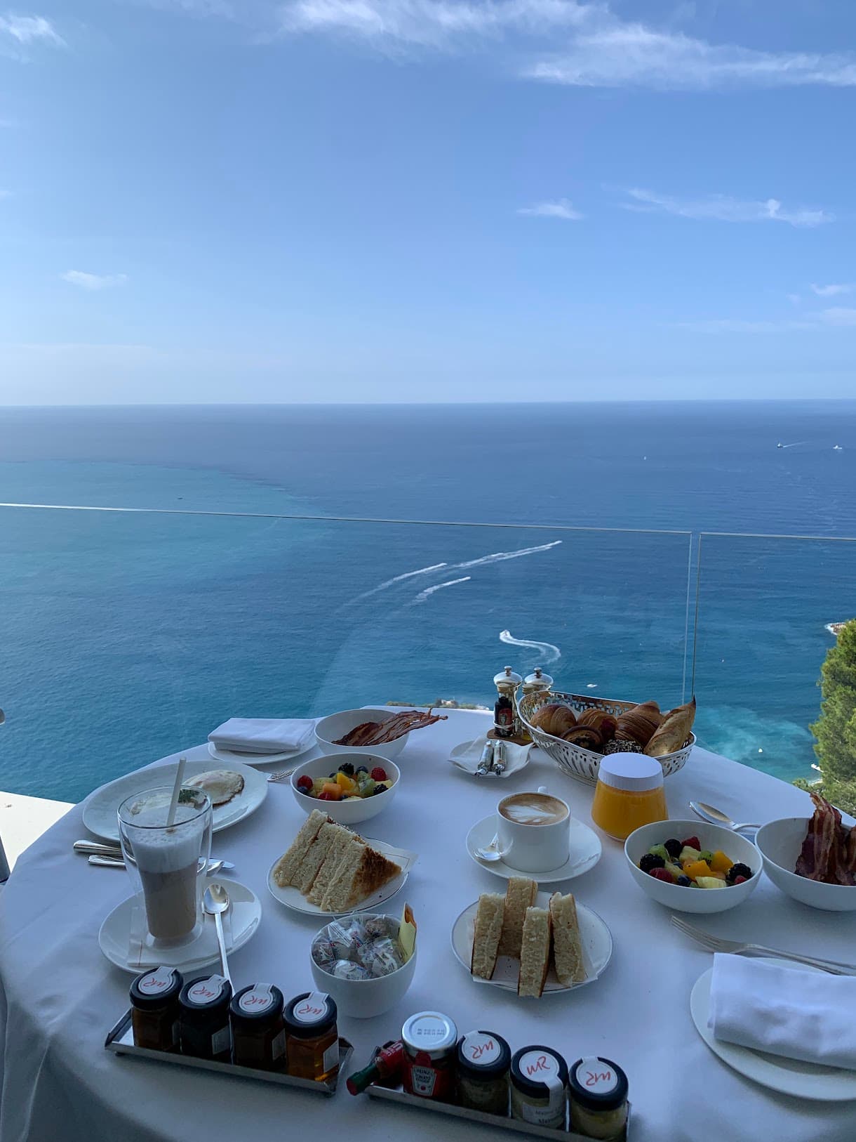 A view of a white table with various treats on top of it, next to a glass ledge that overlooks the blue sea.