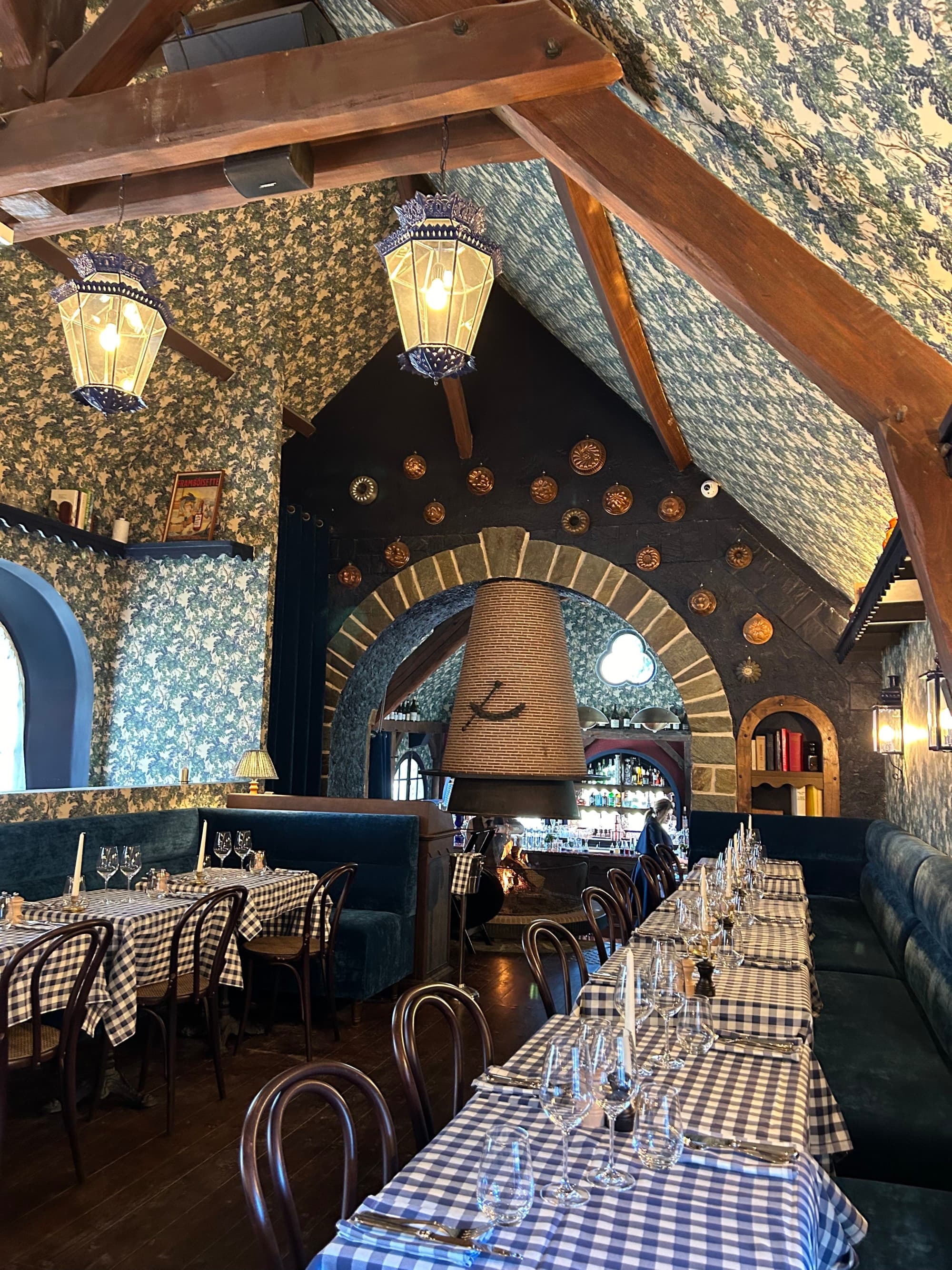 A beautiful dining hall with blue checkered table cloths, wooden chairs, lamp lighting, wood paneled ceilings with patterned wallpaper and a beautiful bar in the background.