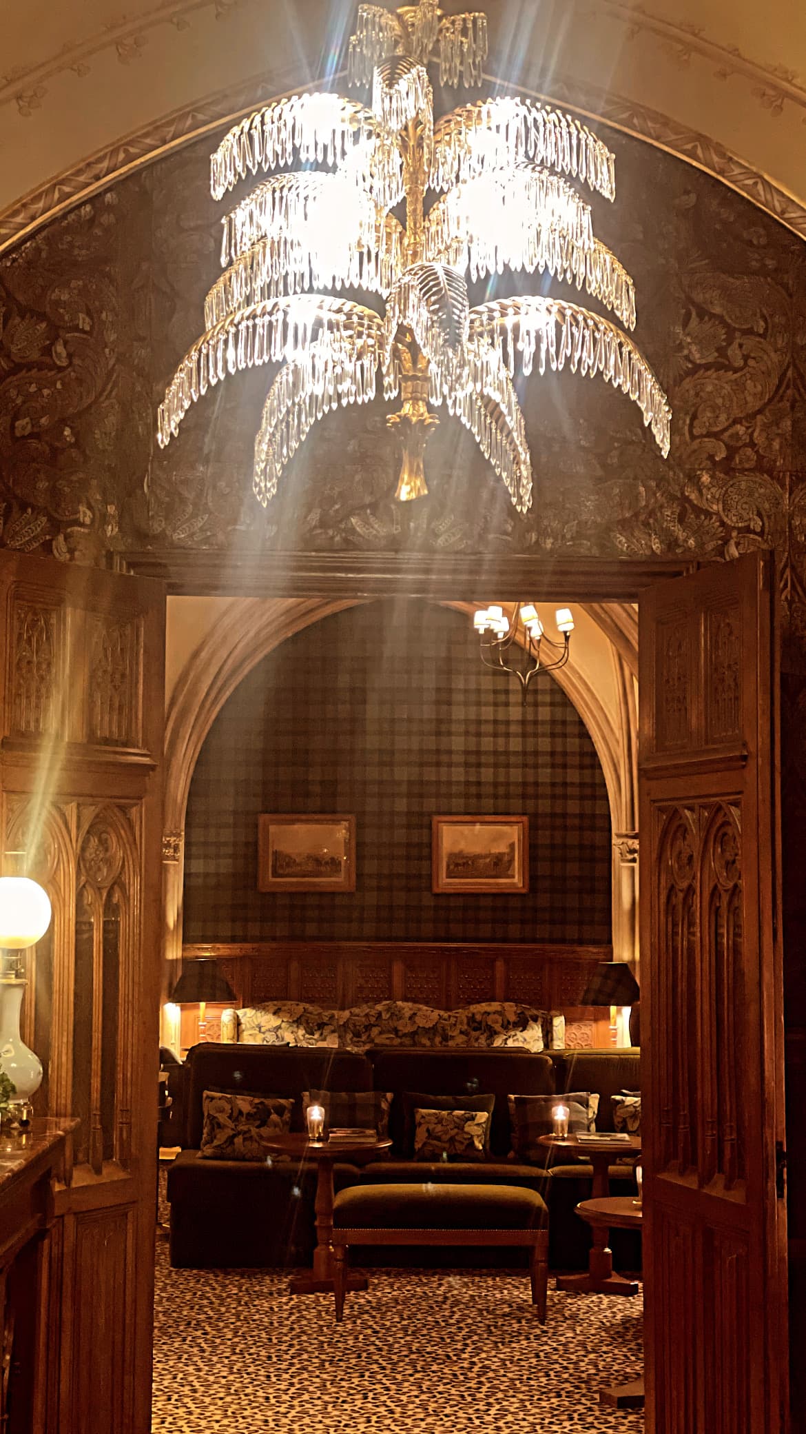 A beautiful art deco chandelier hanging above a doorway with patterned wallpaper that leads to a room with seating arrangements and art hanging on the wall.