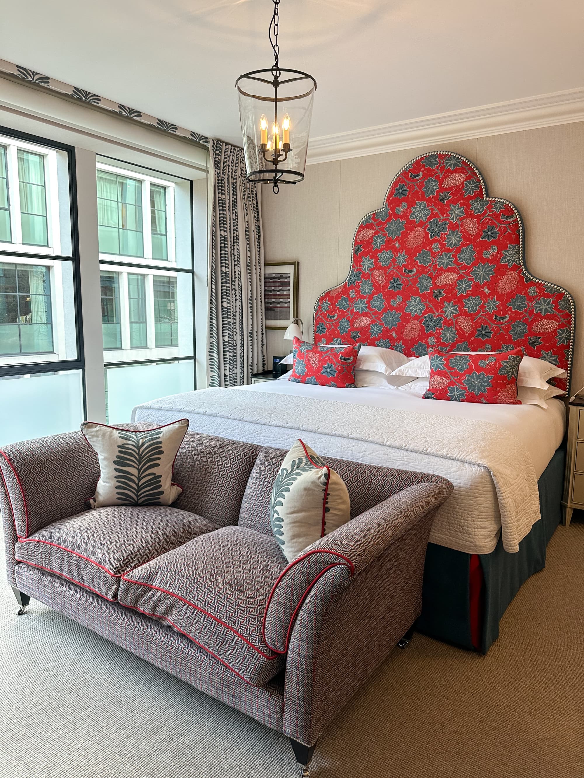 A white bed with a red patterned headboard and matching decorative pillows. There is art on the wall and curtains with a metal chandelier hanging over them. There is also a brown couch with two decorative pillows at the end of the bed.