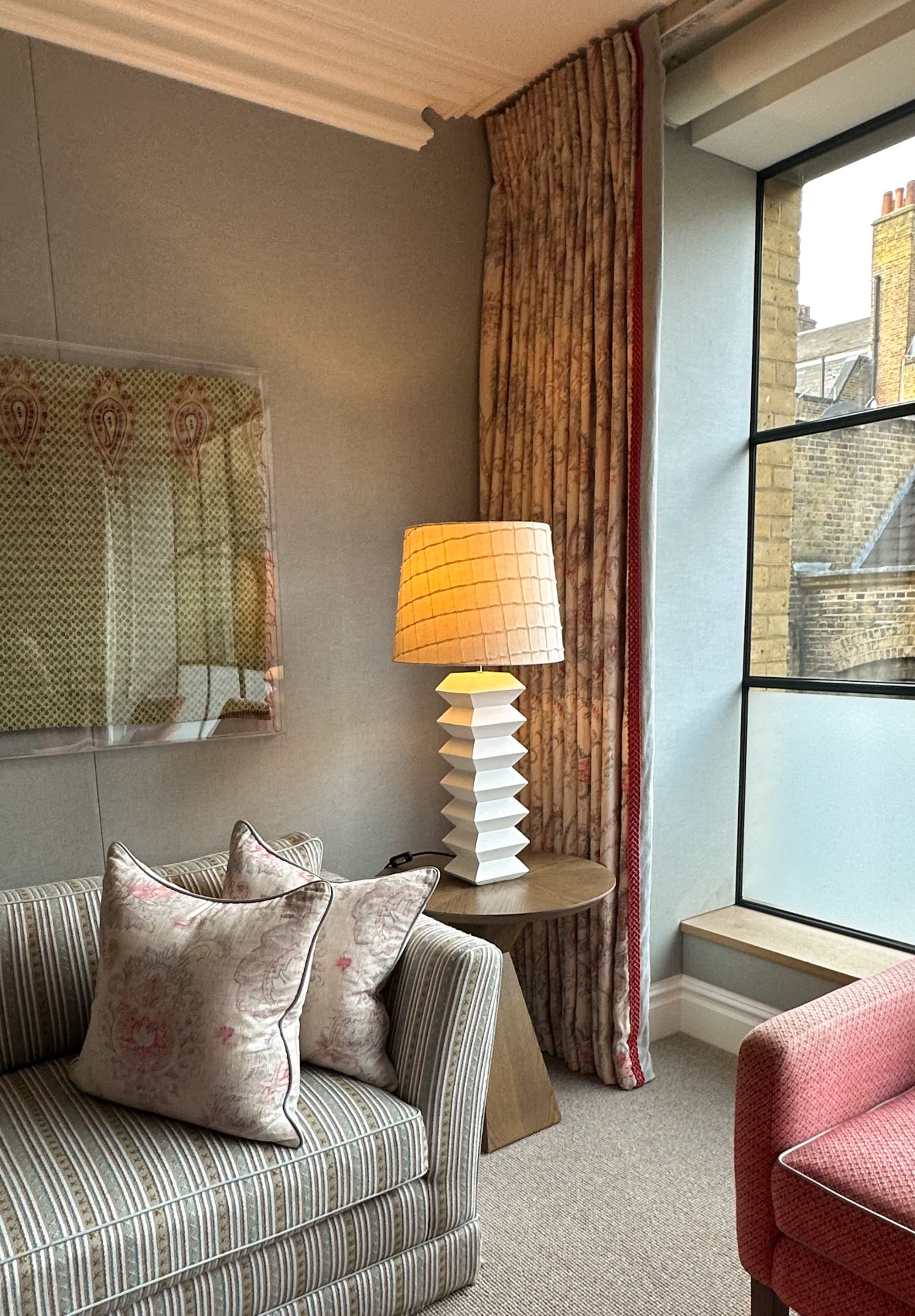 A hotel living room with a patterned sofa, decorative pillows, wall art, table lamp, side table, patterned curtains, windows and red chair on the right hand side of the photo.