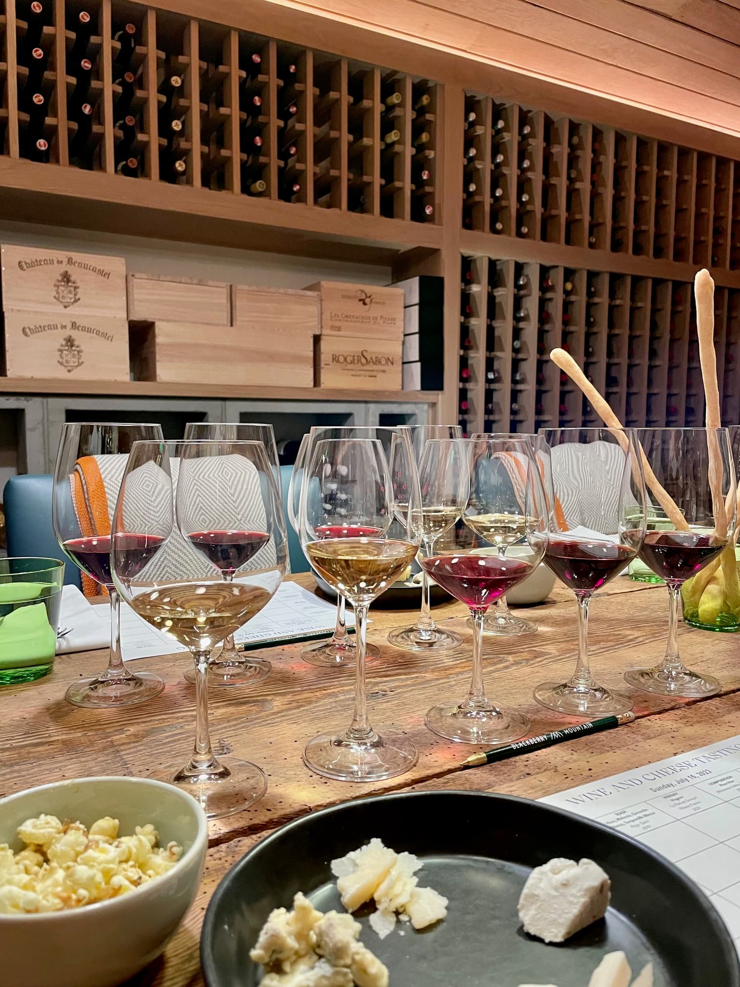 A row of wine glasses, each filled with different kinds. There are also plates of cheese in the front, and wine storage racks in the back.