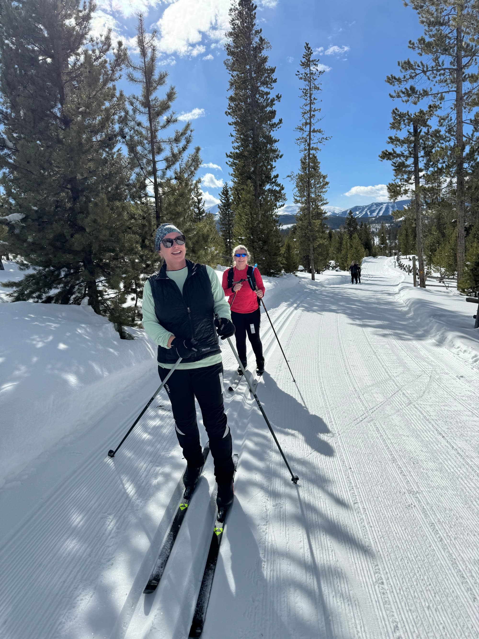 Two people cross country skiing outside with winter gear on. There's snowy terrain and pine trees surrounding them.