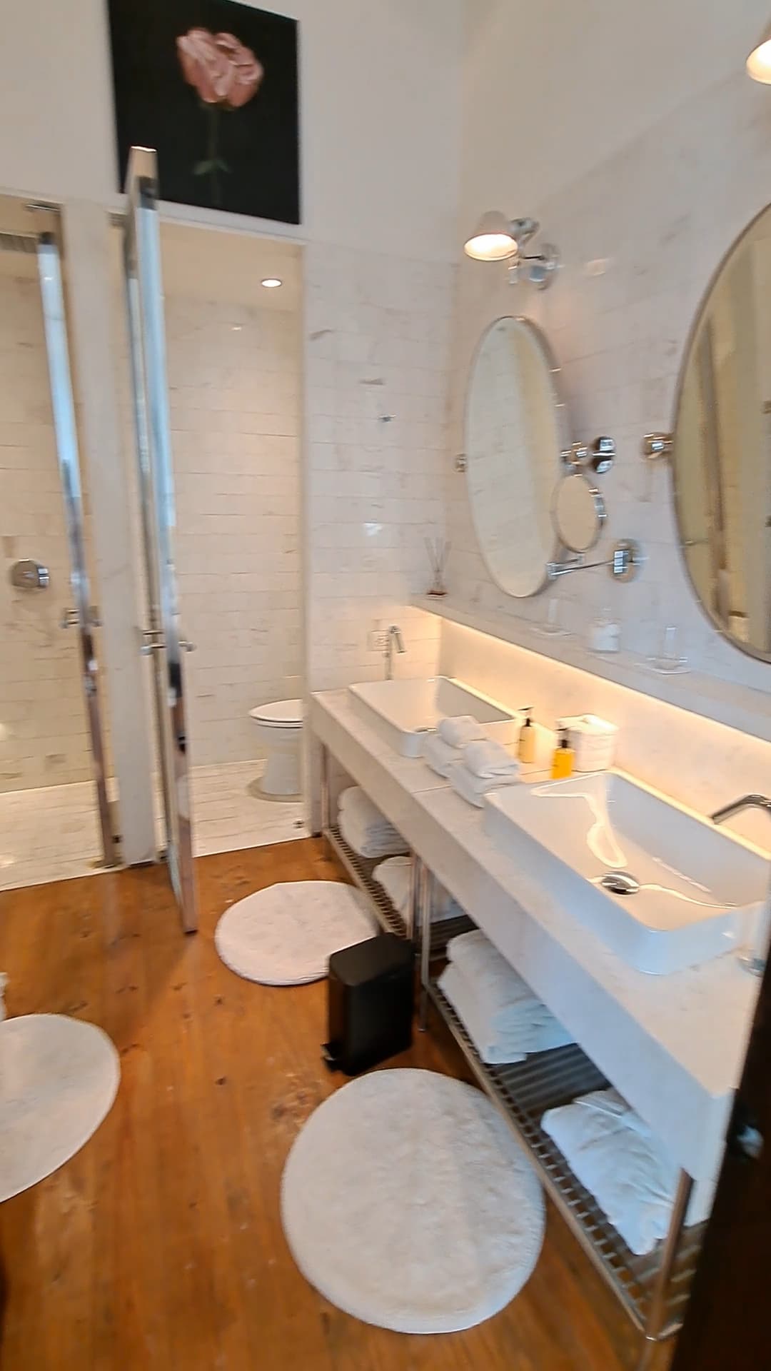 A bathroom with a double sink, three white and round rugs, a shower, round mirrors and pendant lighting. The floor is also wood paneled.
