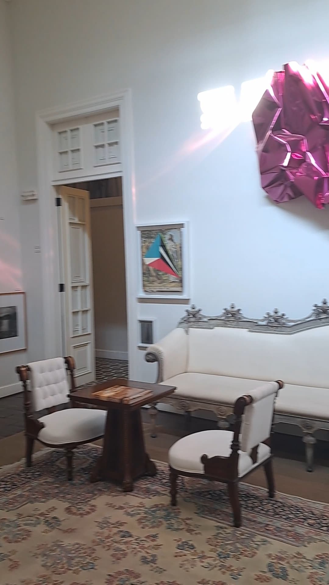 A lounge area complete with a patterned beige rug, two white chairs across from a wooden table, a white sofa and artwork hanging on the wall.