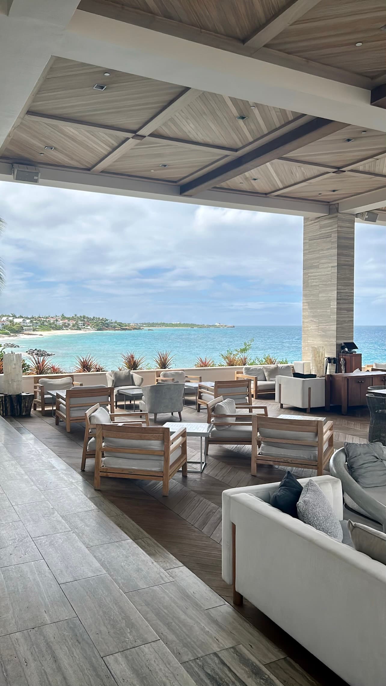 A beautiful lounge area outside complete with white chairs and couches on top of beige tiled flooring and wood beamed ceilings. There is also a stunning view of the light blue water and land in the distance.