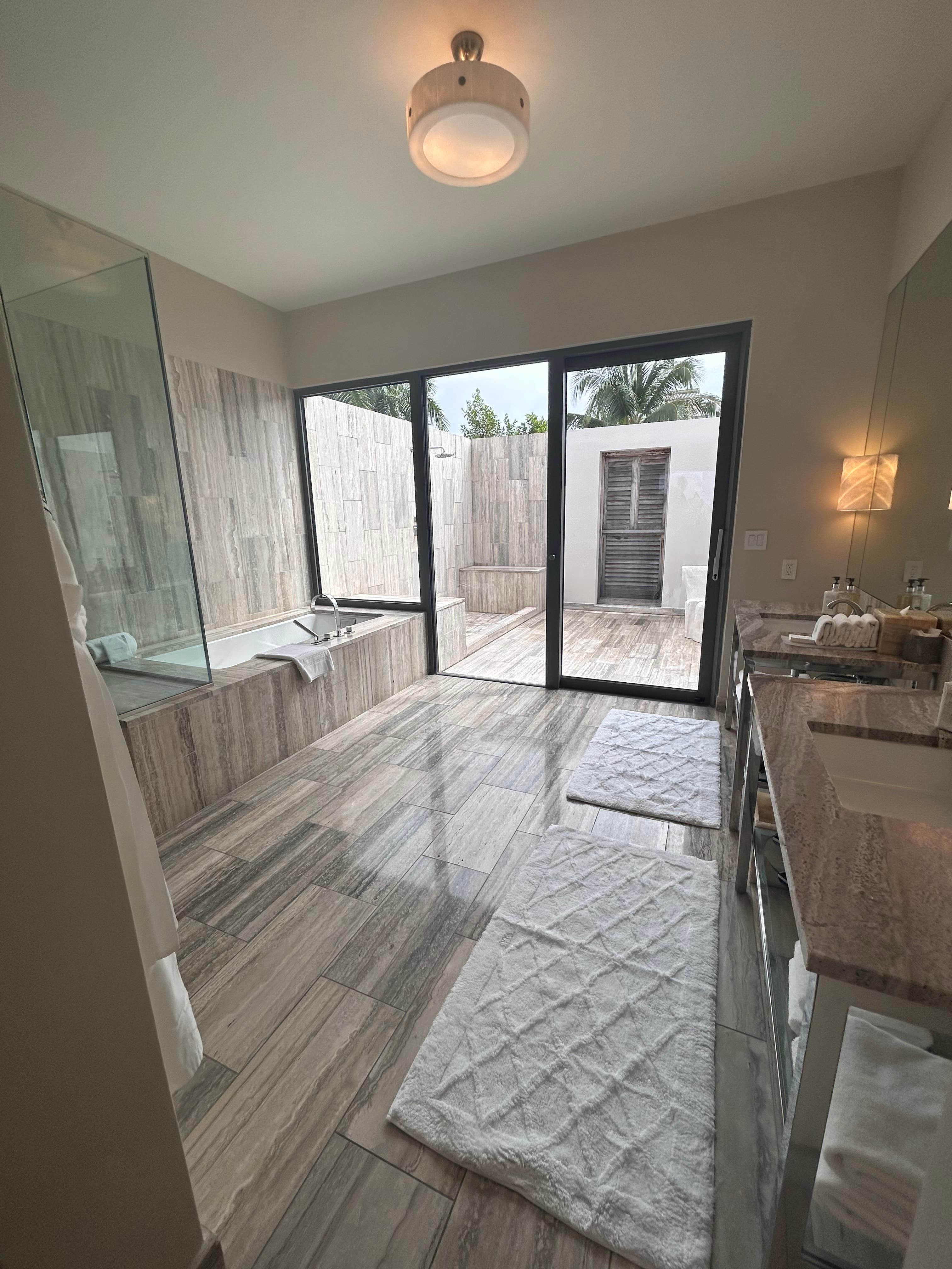 A beautiful hotel suite bathroom complete with beige striped tiling, white rugs, a glass shower next to a large bathtub, glowing sconces and a sliding door leading out to an outdoor shower covered in the same style of tiling.