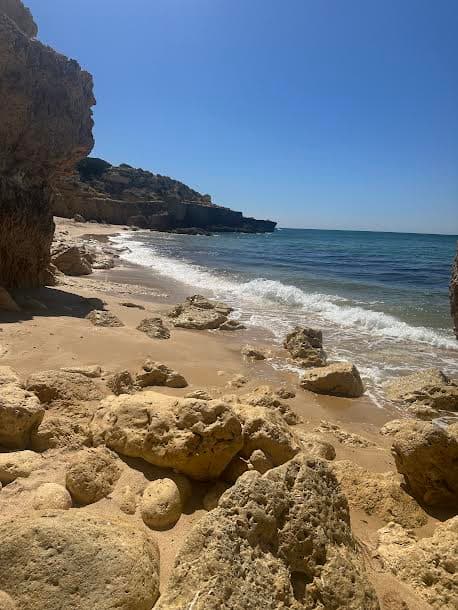 A view of the rocky and sandy beach with small waves rolling in from the blue sea.