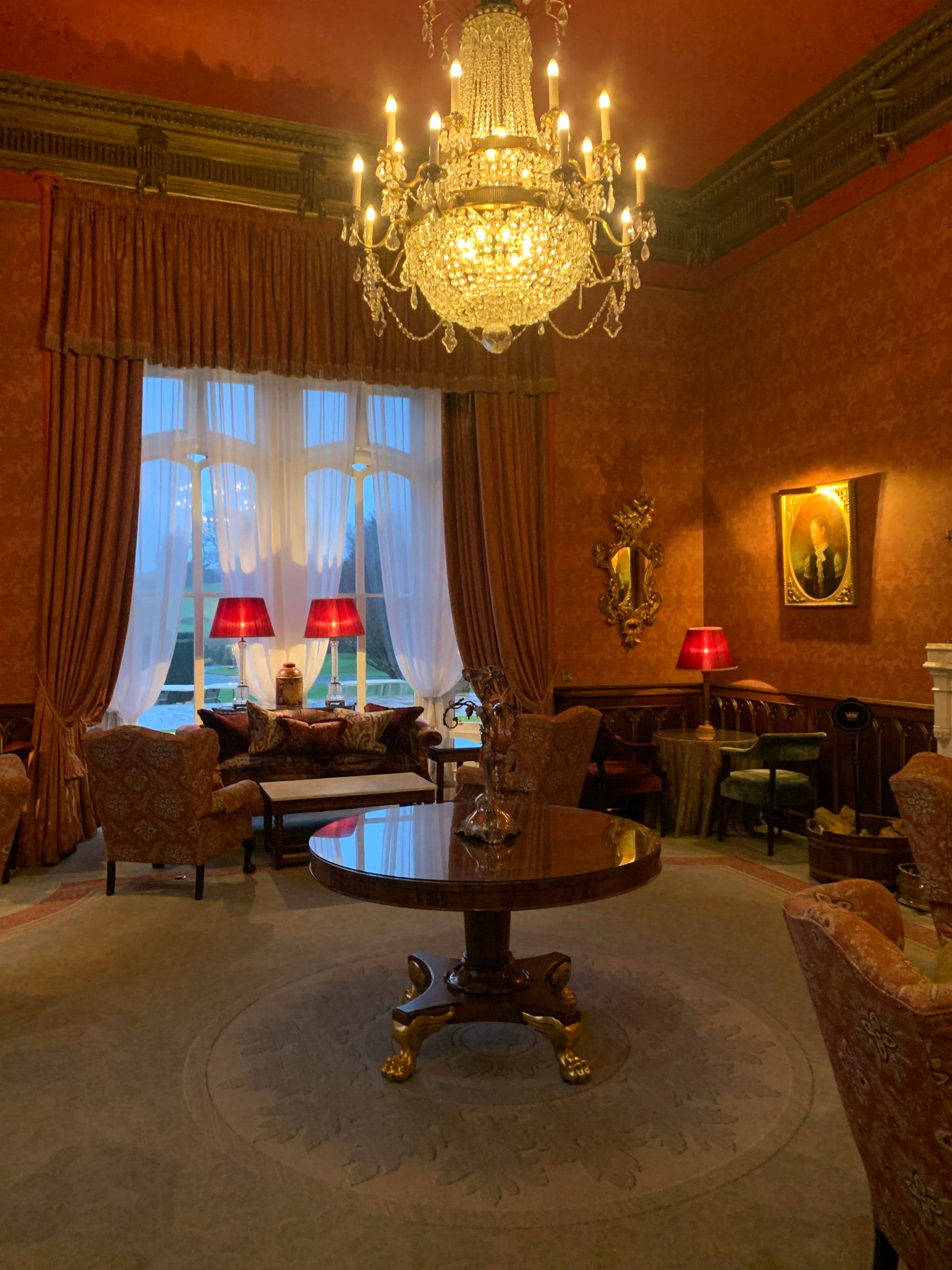 A fancy room complete with a round wooden table, chairs, lamps, artwork and a large crystal chandelier hanging above.