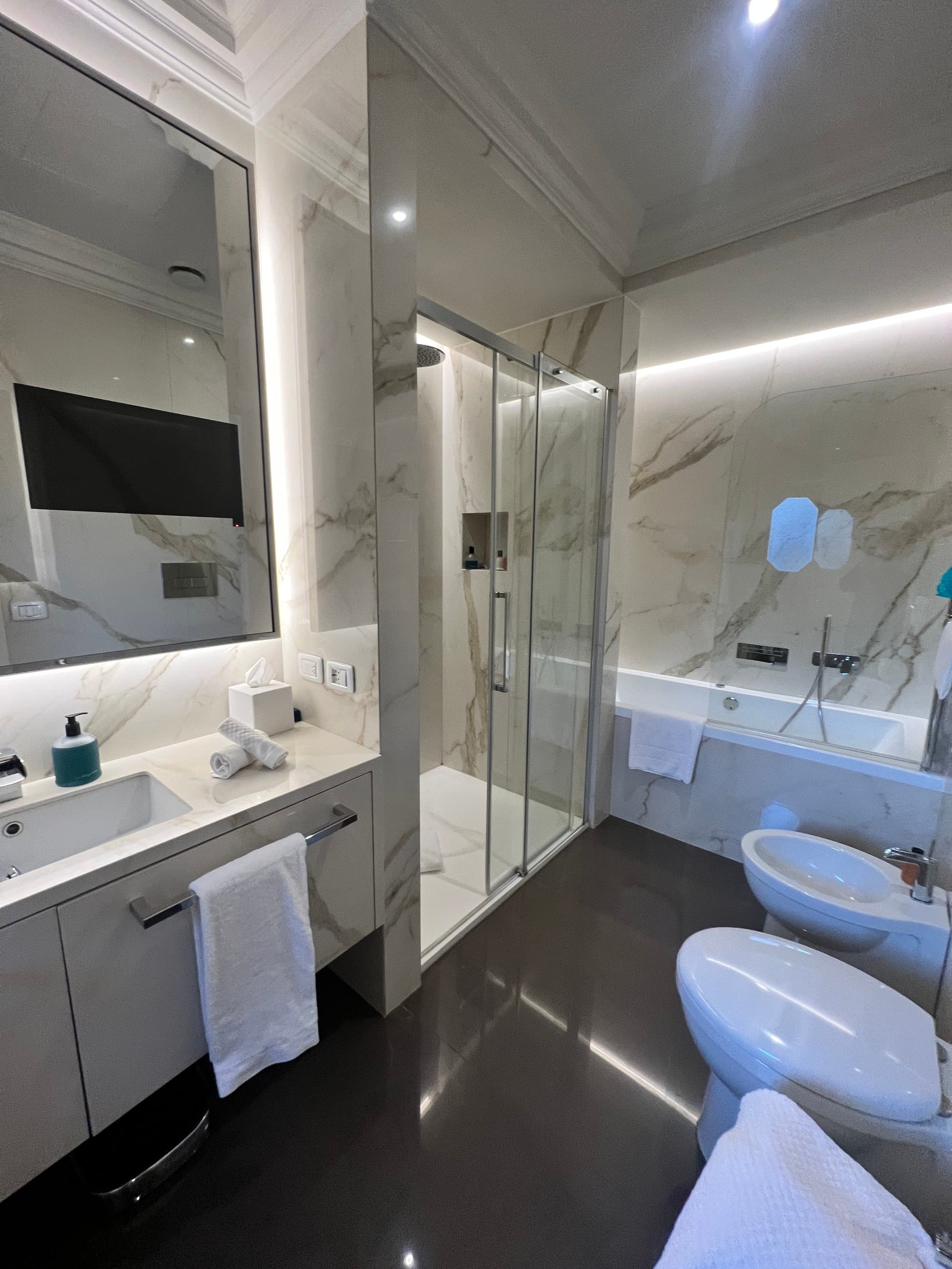 A modern style bathroom with a marble walled bathtub, shower, toilet and lit mirror behind the sink.
