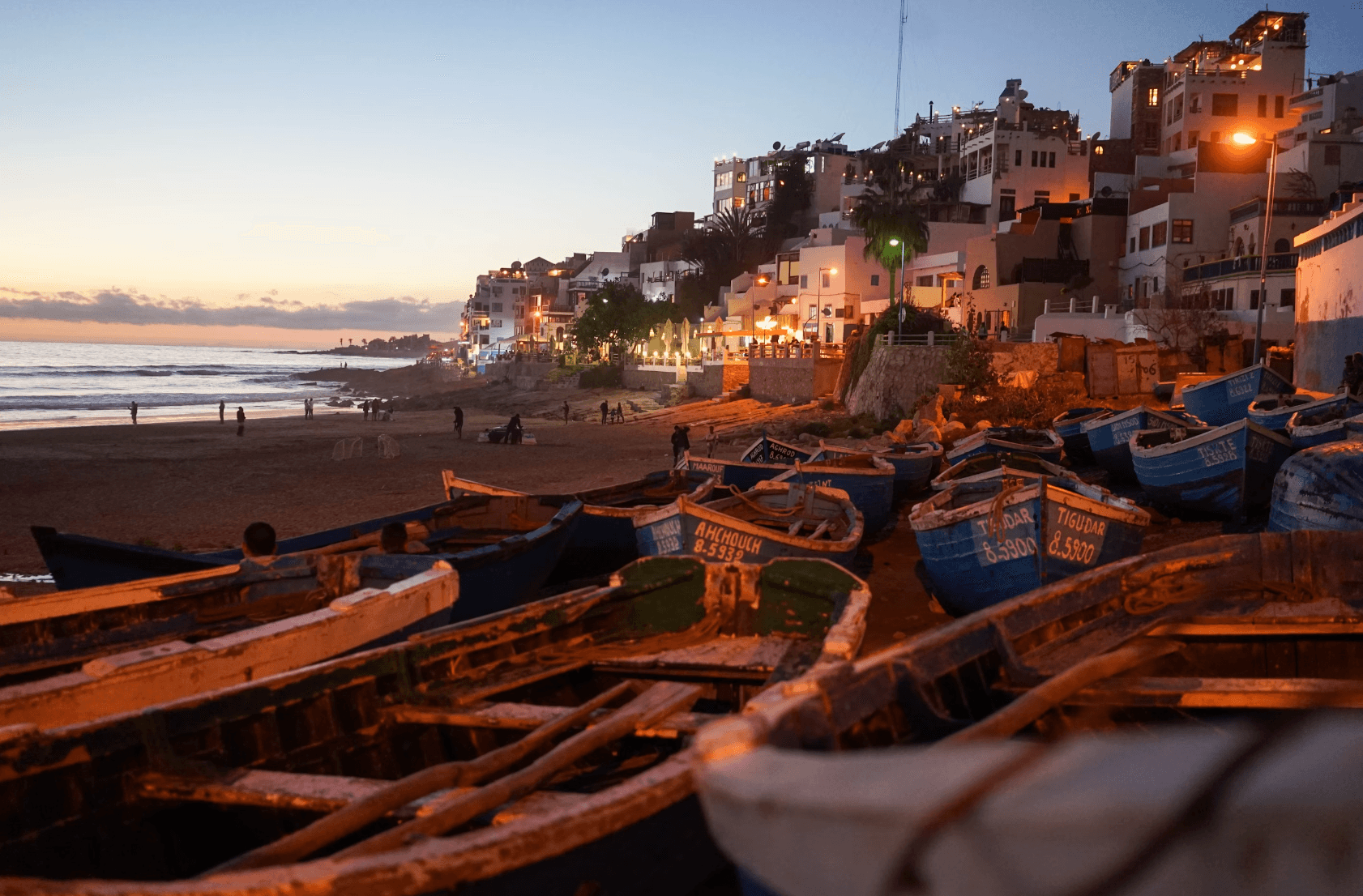 A view of boats on the beach surrounded by sand, buildings in the distance and the sea in the evening. There are people walking along the shoreline and a faint sunset in the distance.