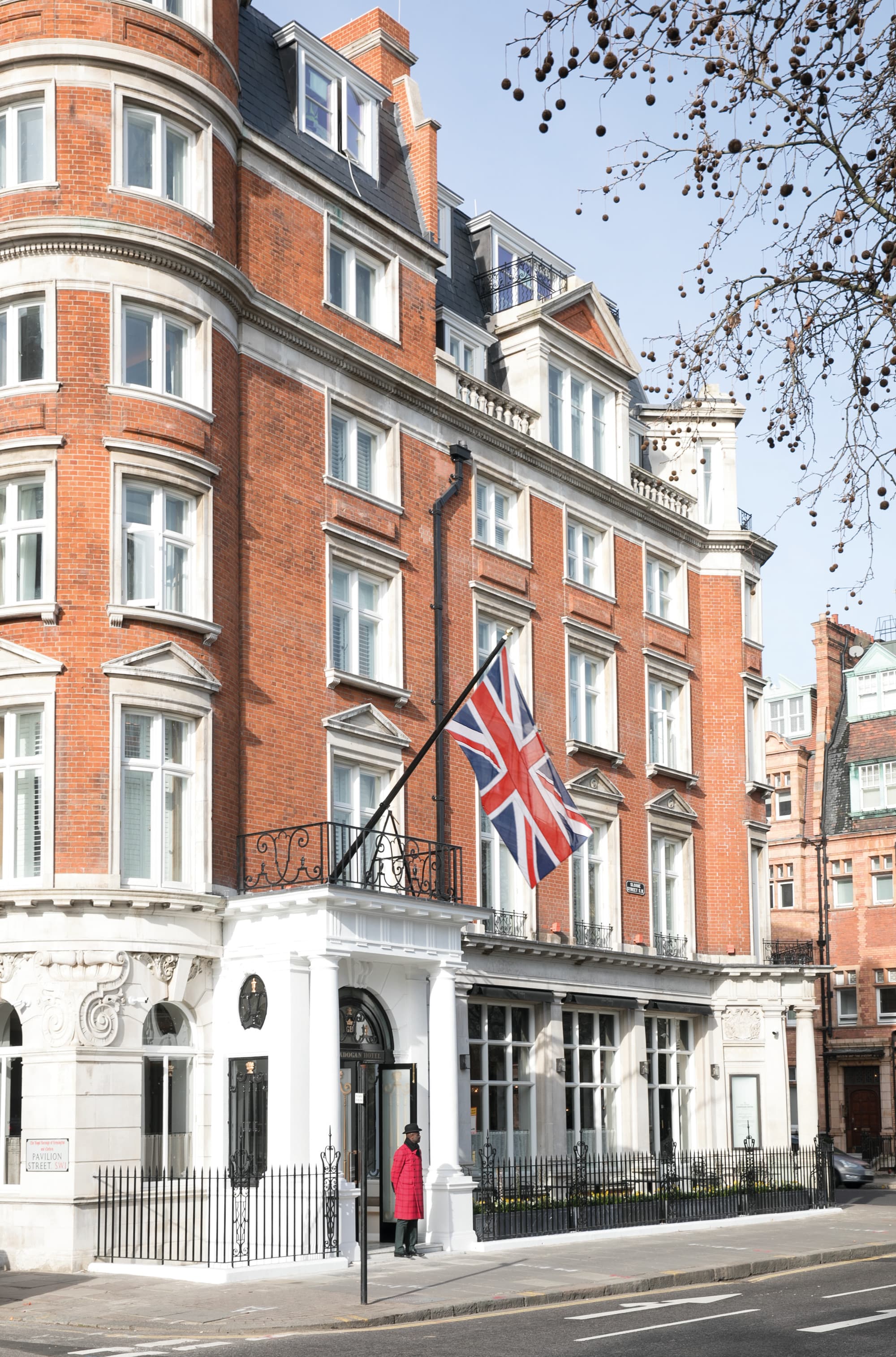 a man in a red coat stands outside of a red-brick building with a British flag above its entrance