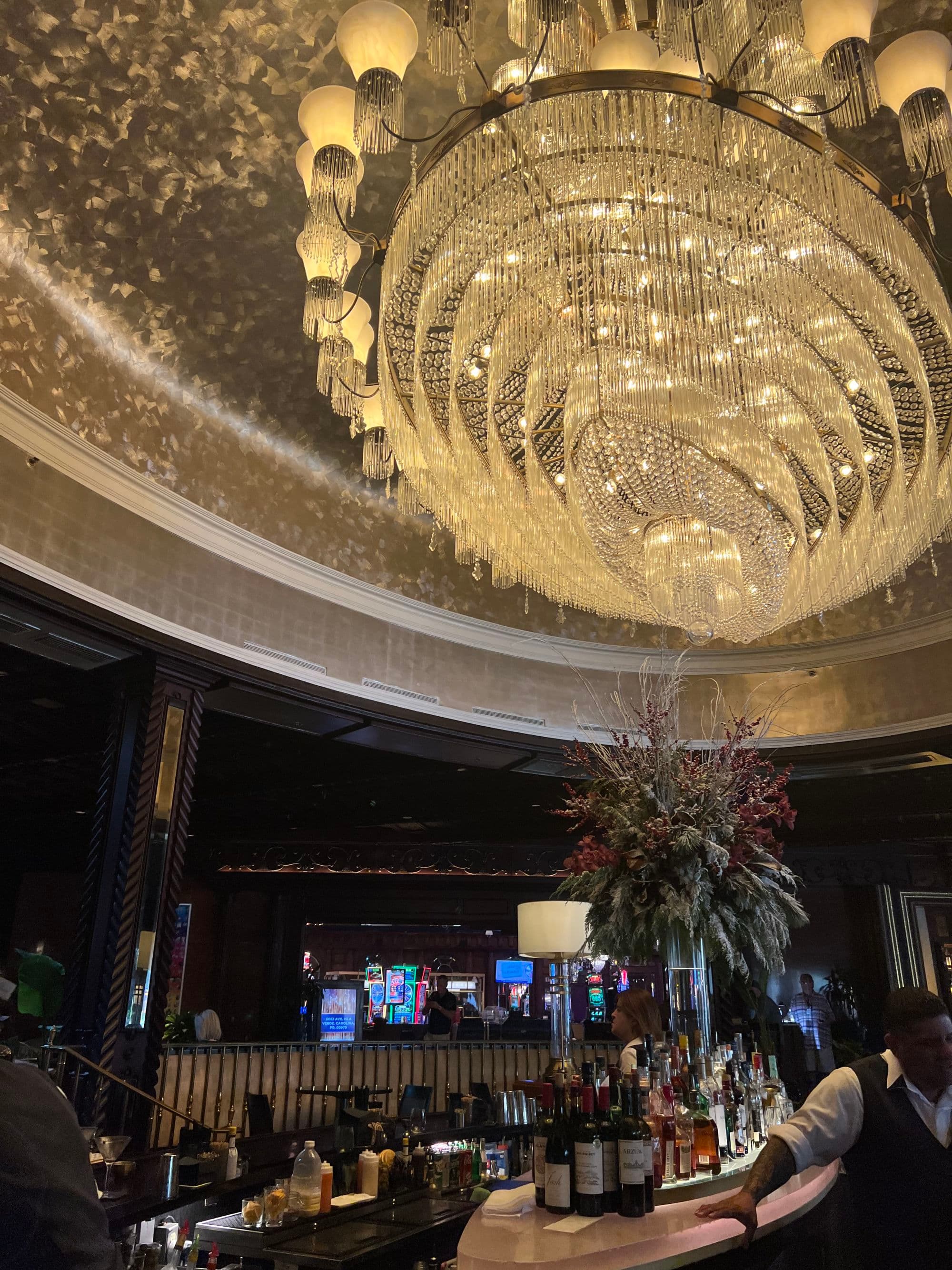 A lobby with a massive crystal and gold chandelier hanging above the bar and people.