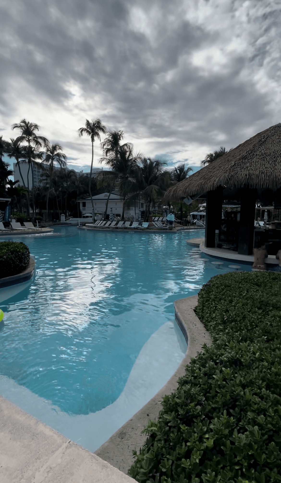 A swimming pool and bar with a straw roof and palm trees in the surrounding areas.