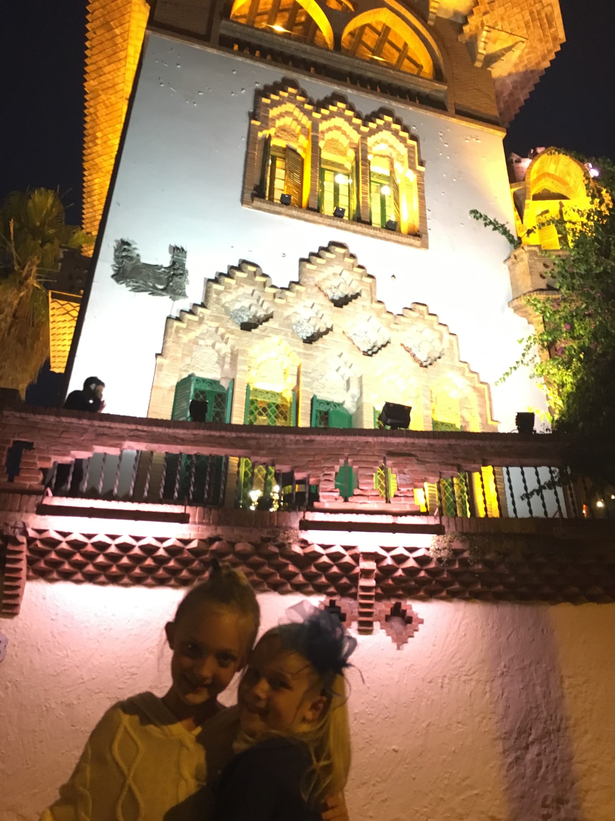 A view of two children smiling in front of a lit up building with intricate window frames, arches and white stone.