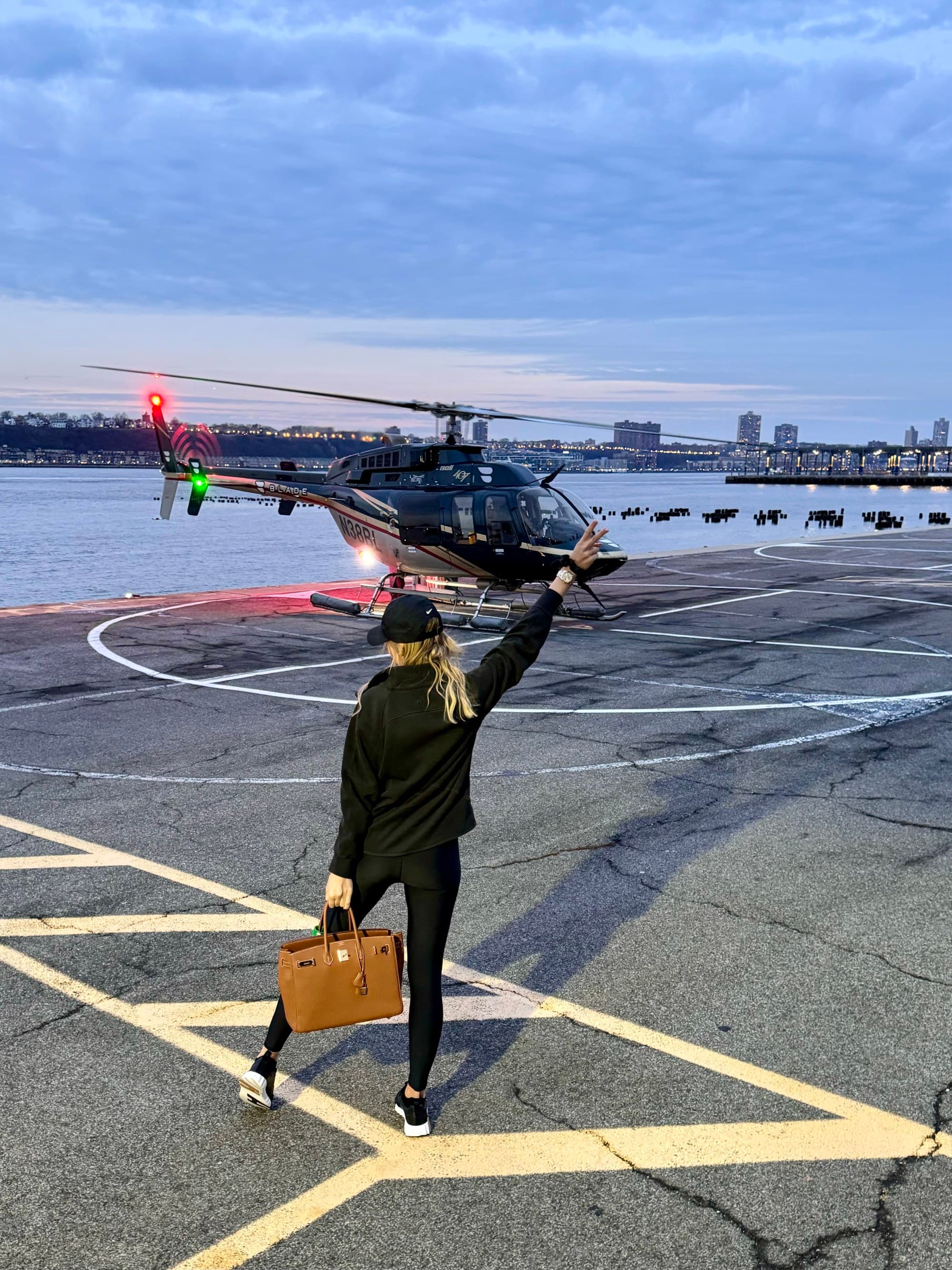 A view of Agnese posing with their hand in the air in front of a helicopter with water and a skyline view in the background at dusk.