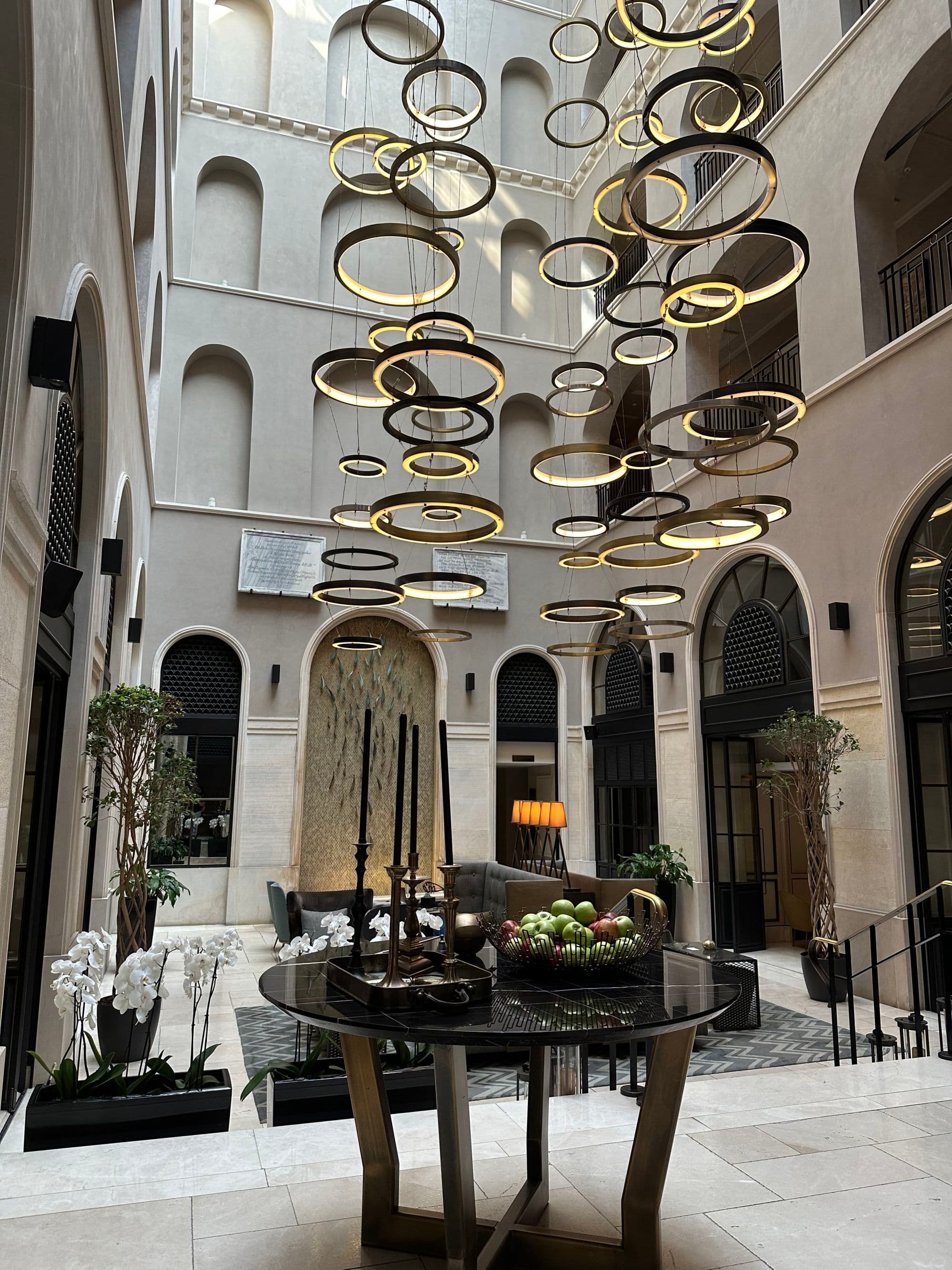 An indoor courtyard with natural light coming from above and modern circular chandeliers in the center above a table with a fruit bowl on top