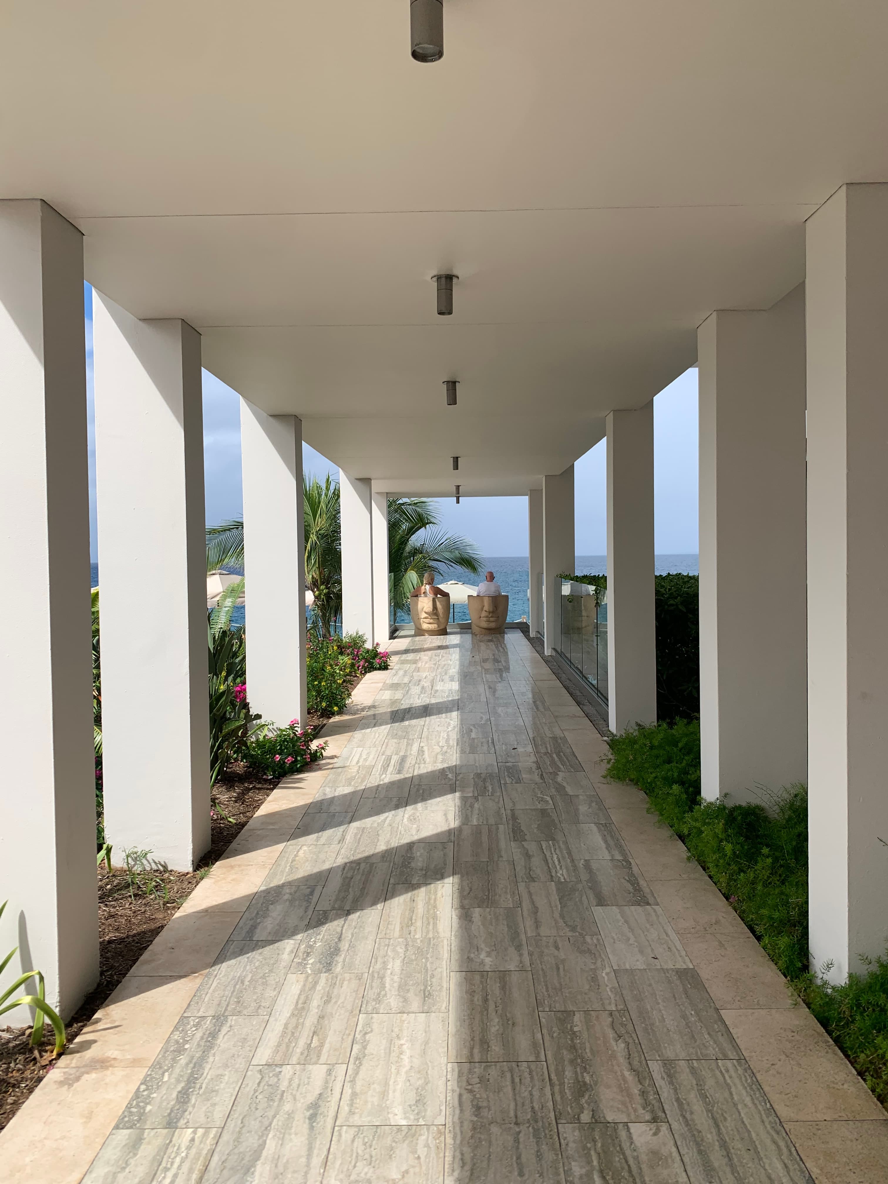 A beige tiled pathway with white pillars and beautiful plants leading to a view of the blue sea.