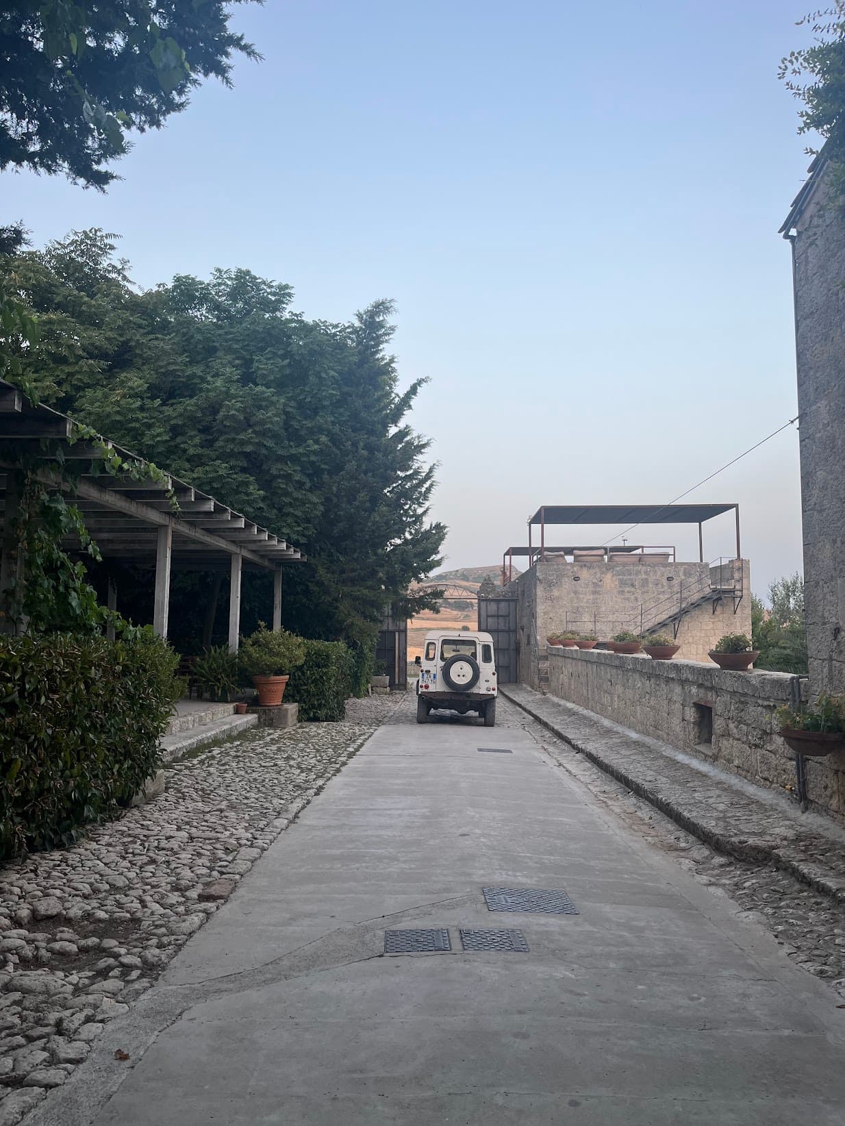 A driveway surrounded by stones, bushes, trees and potted plants. There is a car at the end of the driveway, and more sculptural features in the surrounding areas.