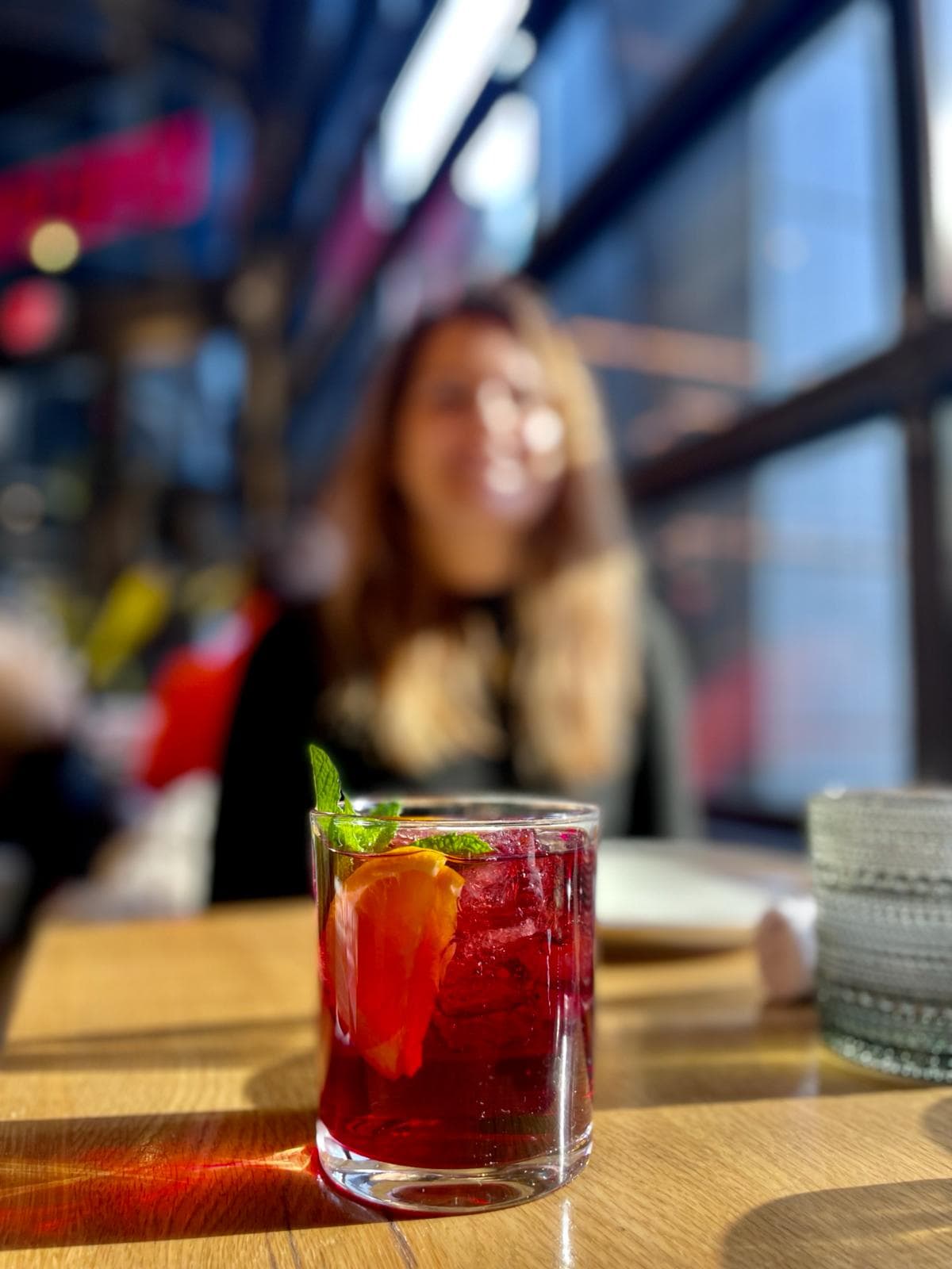 A red cocktail placed on top of a wooden table with a person blurred out in the background.