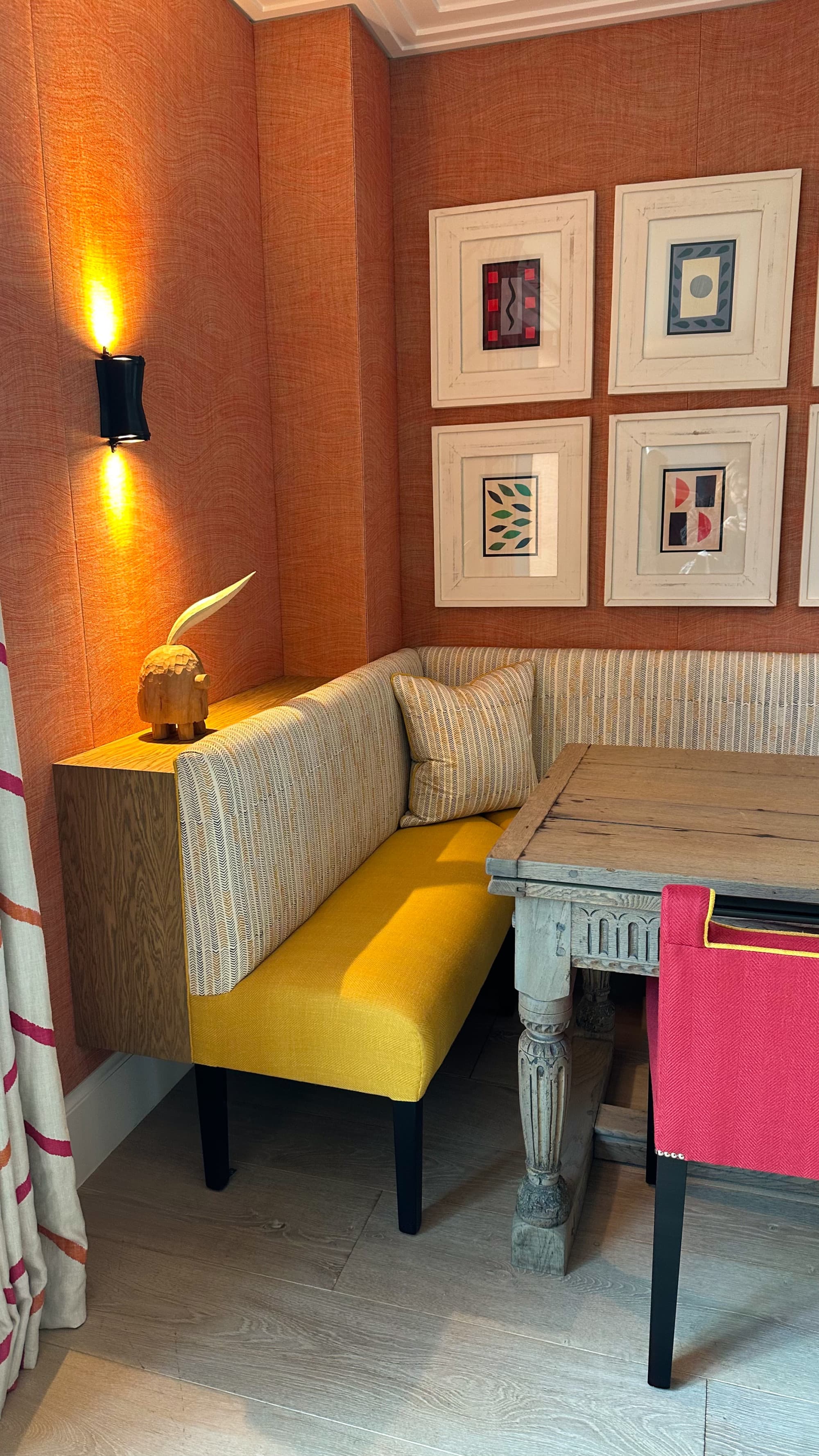 A hotel nook complete with an orange wall, lit sconce, four pieces of art hung, a table, pink chair, gold and patterned banquette and patterned curtain on the left hand side of the photo.