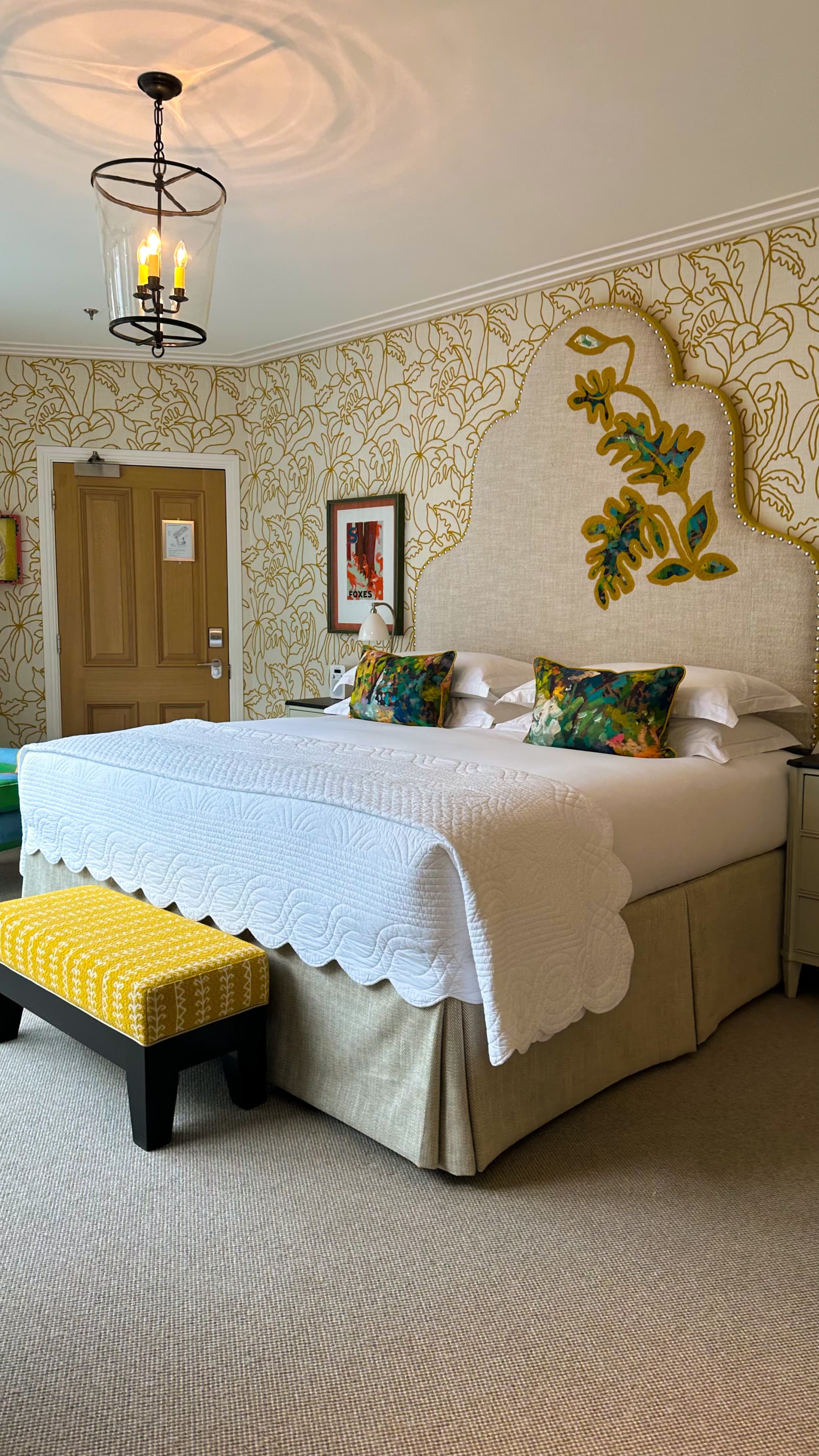 A hotel bedroom with a luxurious white bed next to a tall, beige headboard with a leaf design, patterned wallpaper, yellow bench and door beneath a metal chandelier.