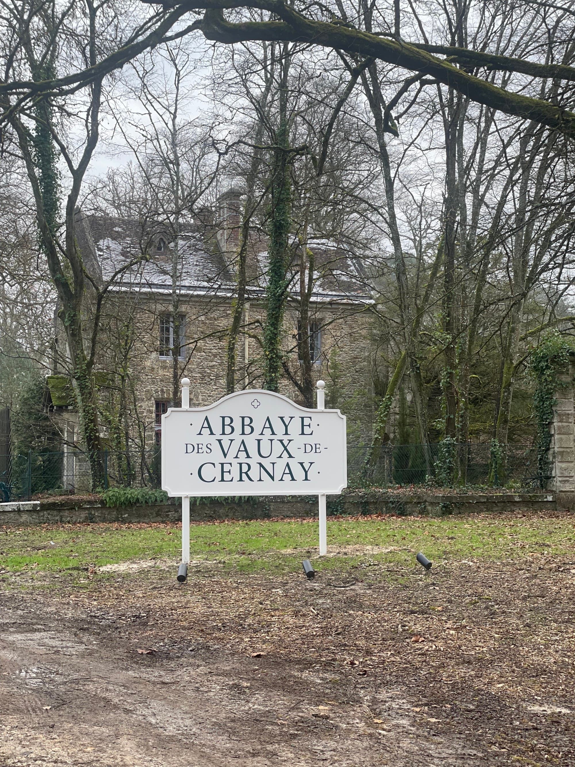 A white sign positioned on top of the grass and dirt road with trees and a stone building in the background that reads "Abbaye des Vaux de Cernay".