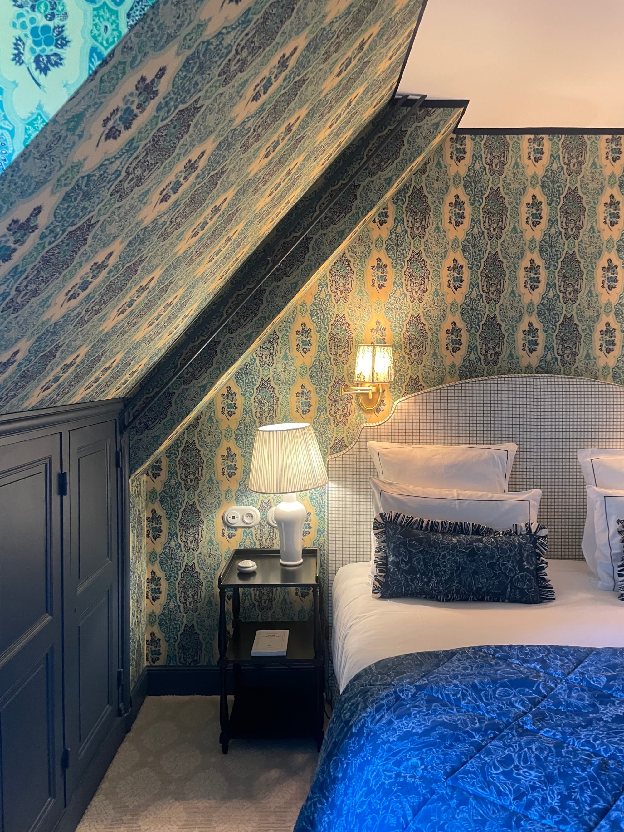 A beautiful hotel bedroom with patterned and blue-toned wallpaper, a white and blue bed, dark side table, white table lamp and sconce lighting.