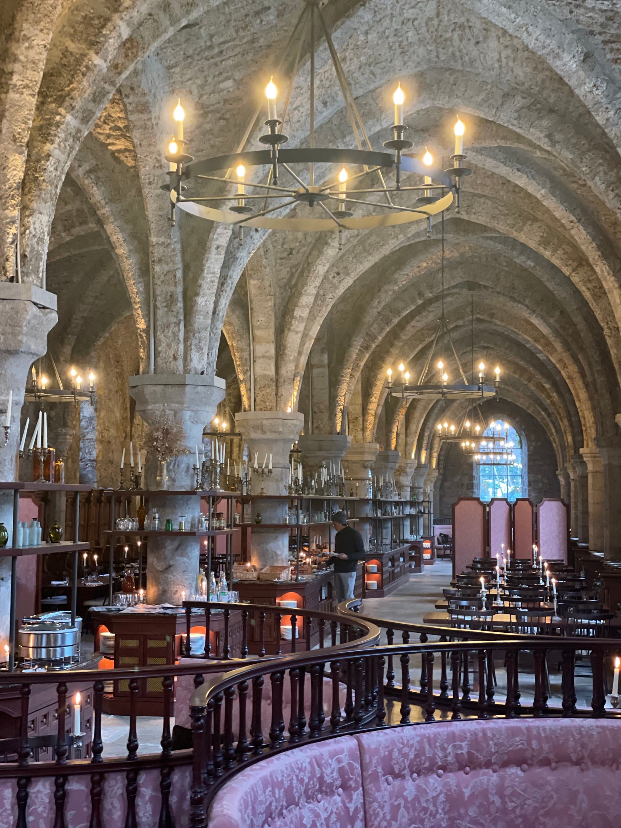 A beautiful stone walled room with arched ceilings, grand metal chandeliers and wood detailed seating areas.