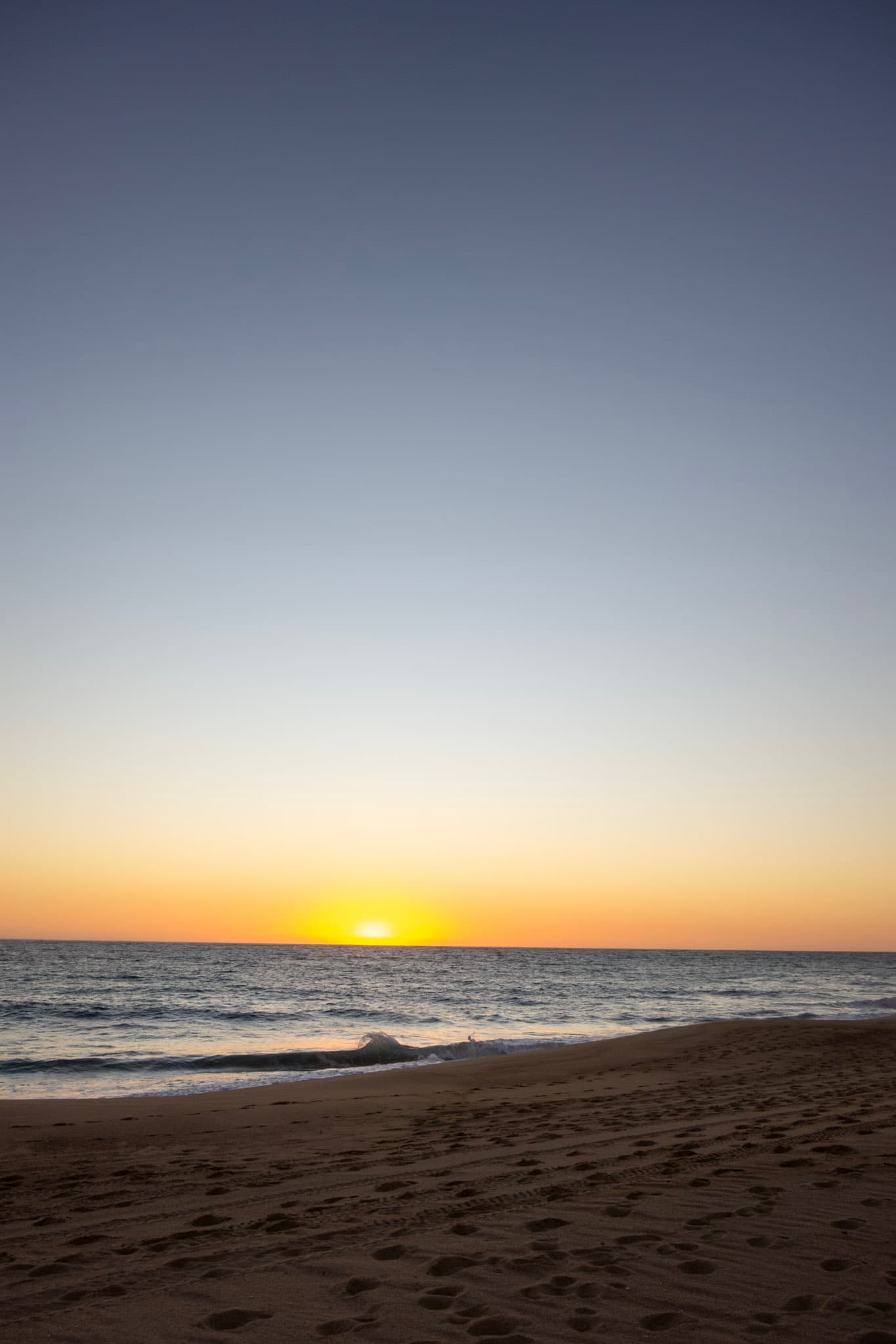 A picture of a beach and the rippling blue sea water with a golden sunset shining from above.