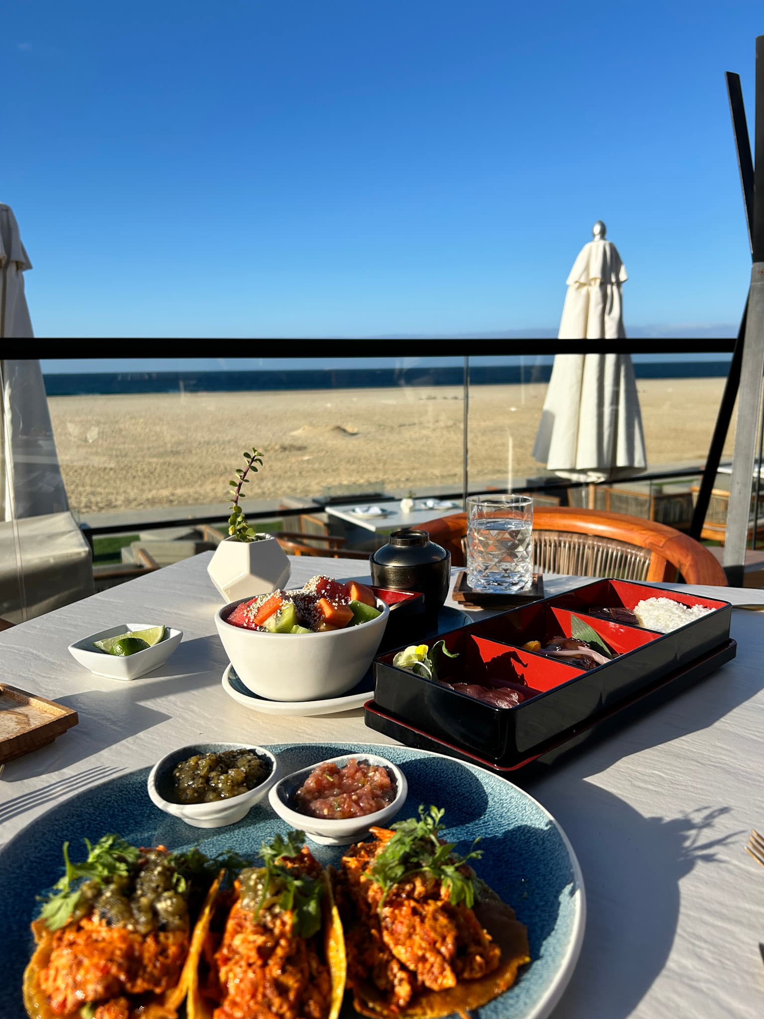 A view of a table with colorful food on top of it. There is a glass barricade in the background looking over the beach and white umbrellas.