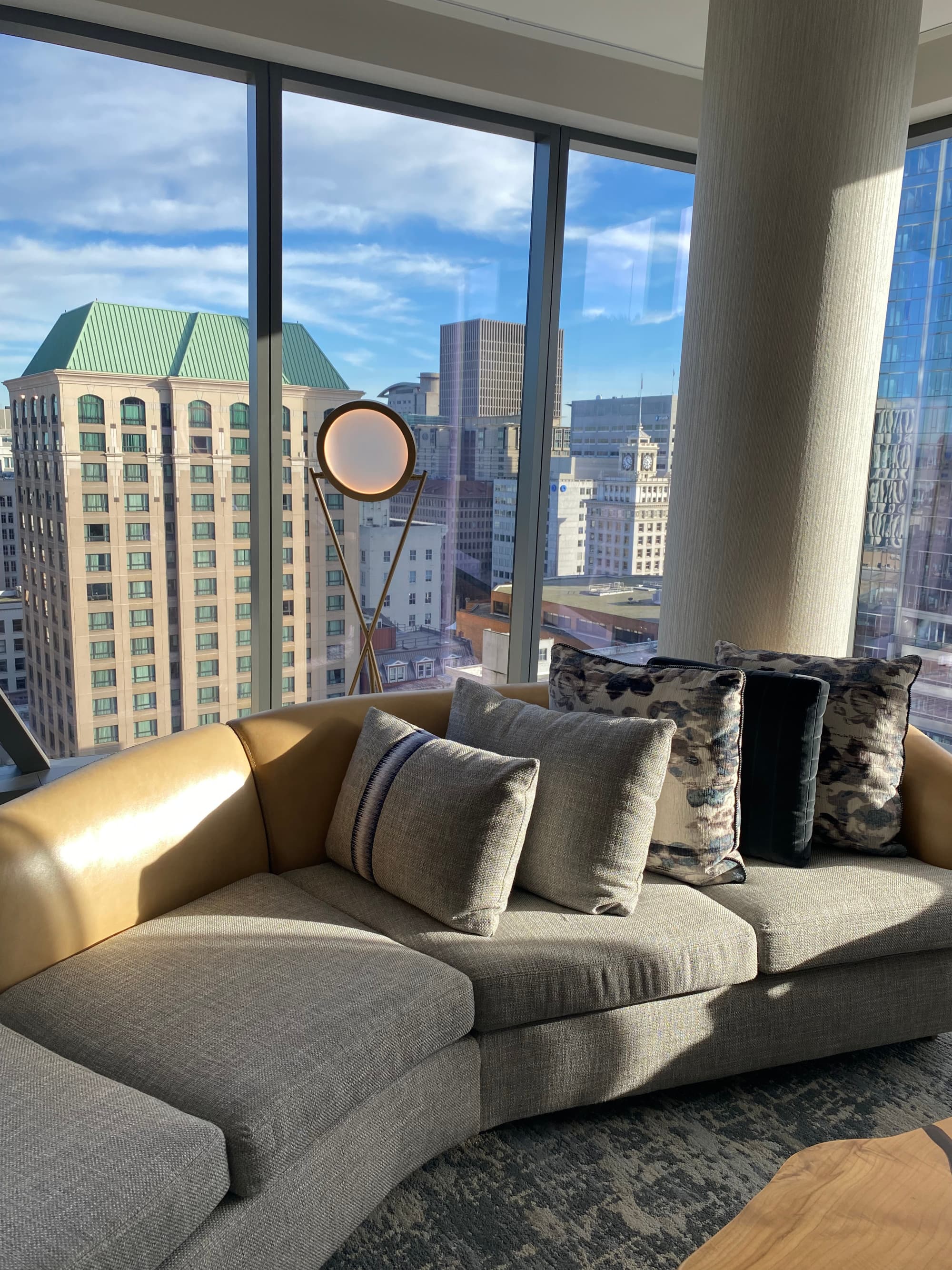 A hotel suite with a modern sofa, decorative pillows and a round mirror next to a large white pillar. There is a view of a city outside of the floor to ceiling windows.