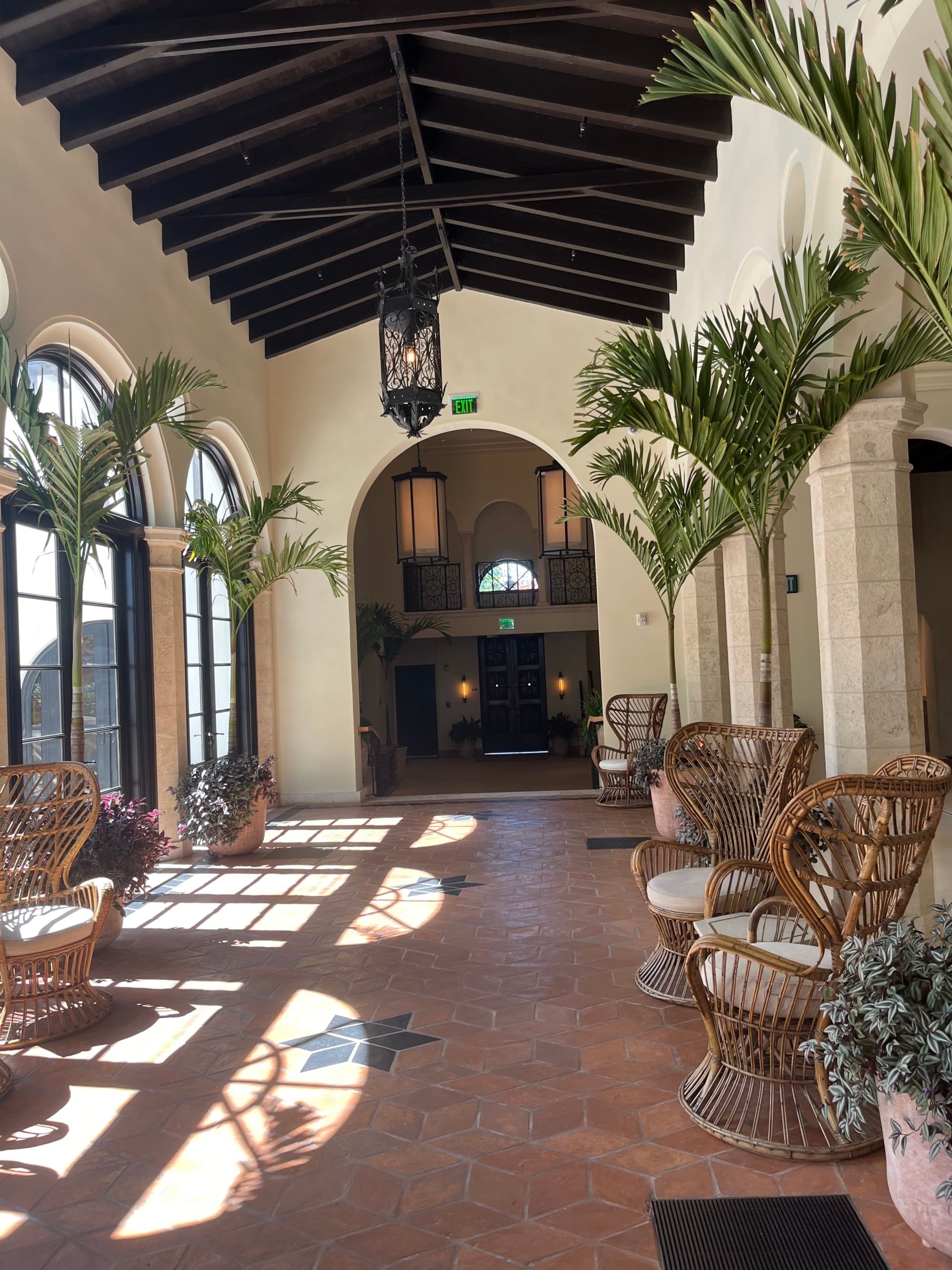 A hotel lobby with wicker chairs, arched windows, palm trees, wood paneled ceilings and a beautiful hanging chandelier.
