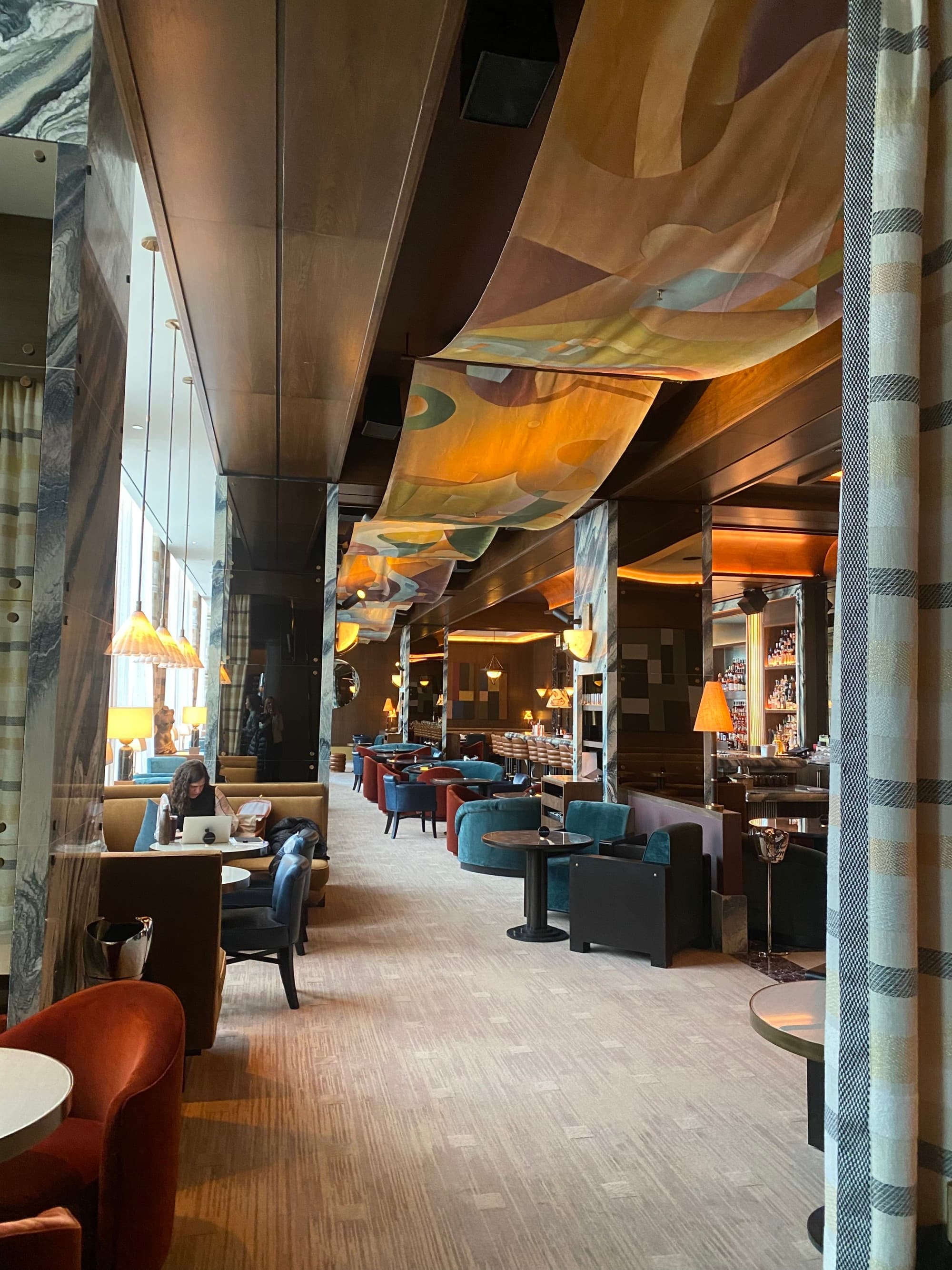 A luxurious hotel lounge with beige carpeting, various velvet chairs and tables, large floor to ceiling windows and gold artwork draped along the ceiling. There are people dining and soft ambient lighting during the daytime.