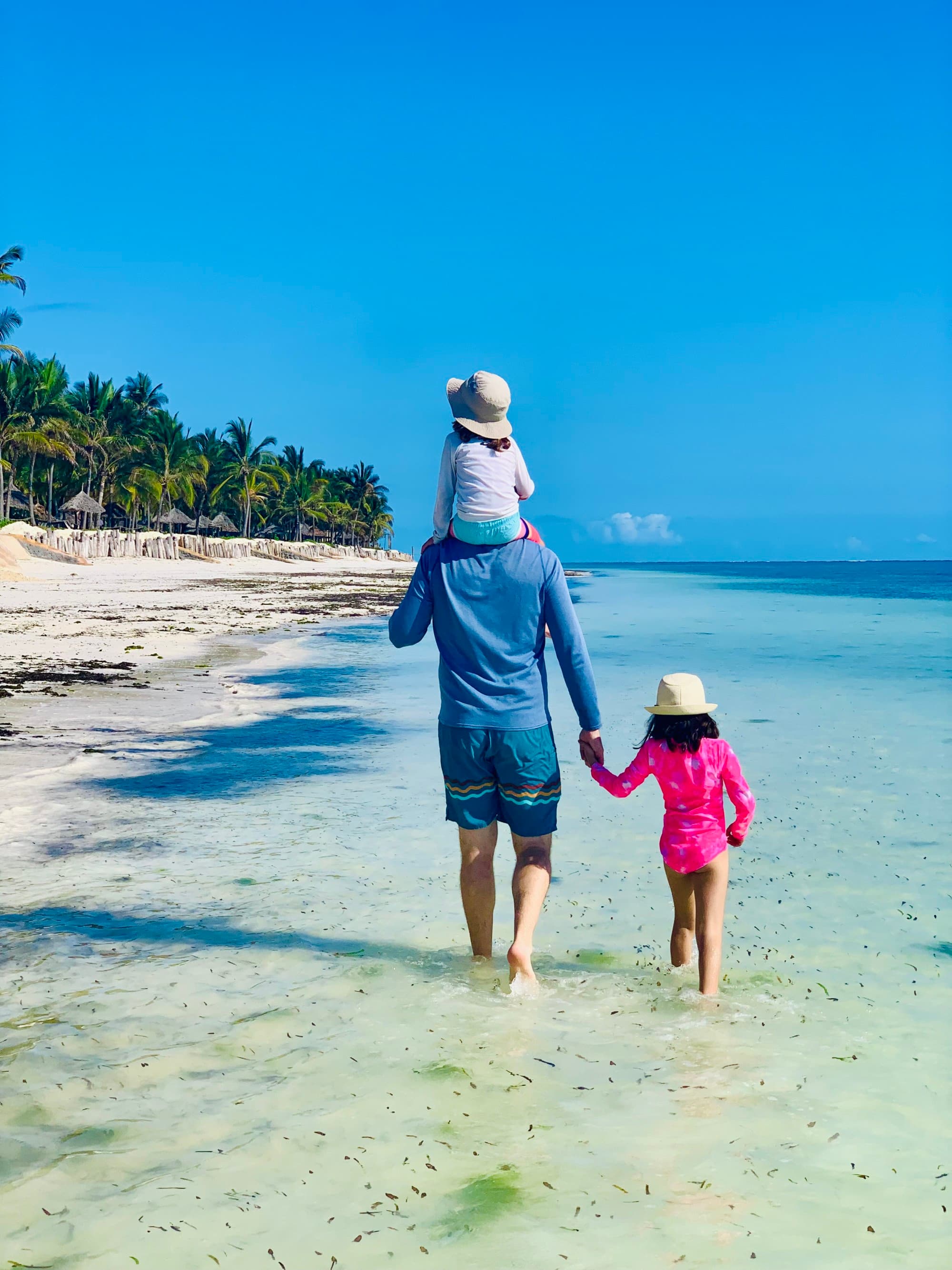 a man in a blue shirt carries a child on his shoulders and holds the hand of another child as the trio walk in turquoise water on a beach