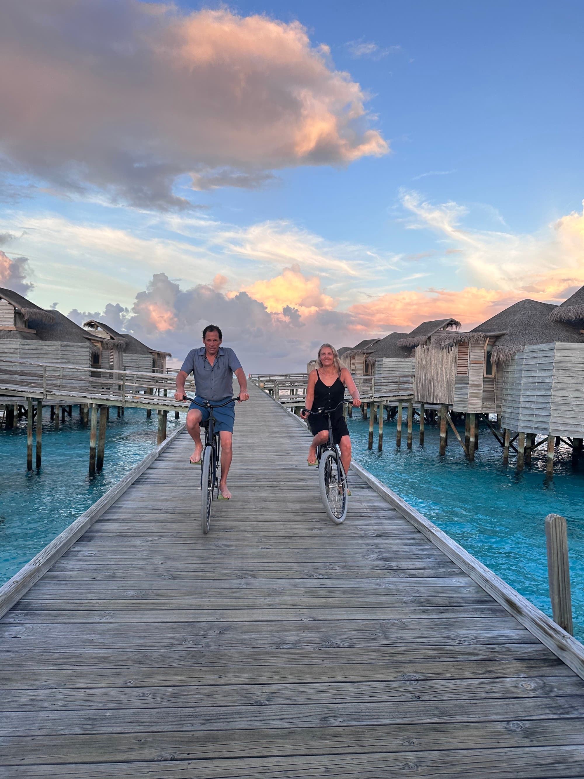 Two people cycling down a wooden bridge surrounded by beach bungalows and a beautiful cloudy sky. There is turquoise blue water on each side of the bridge too.