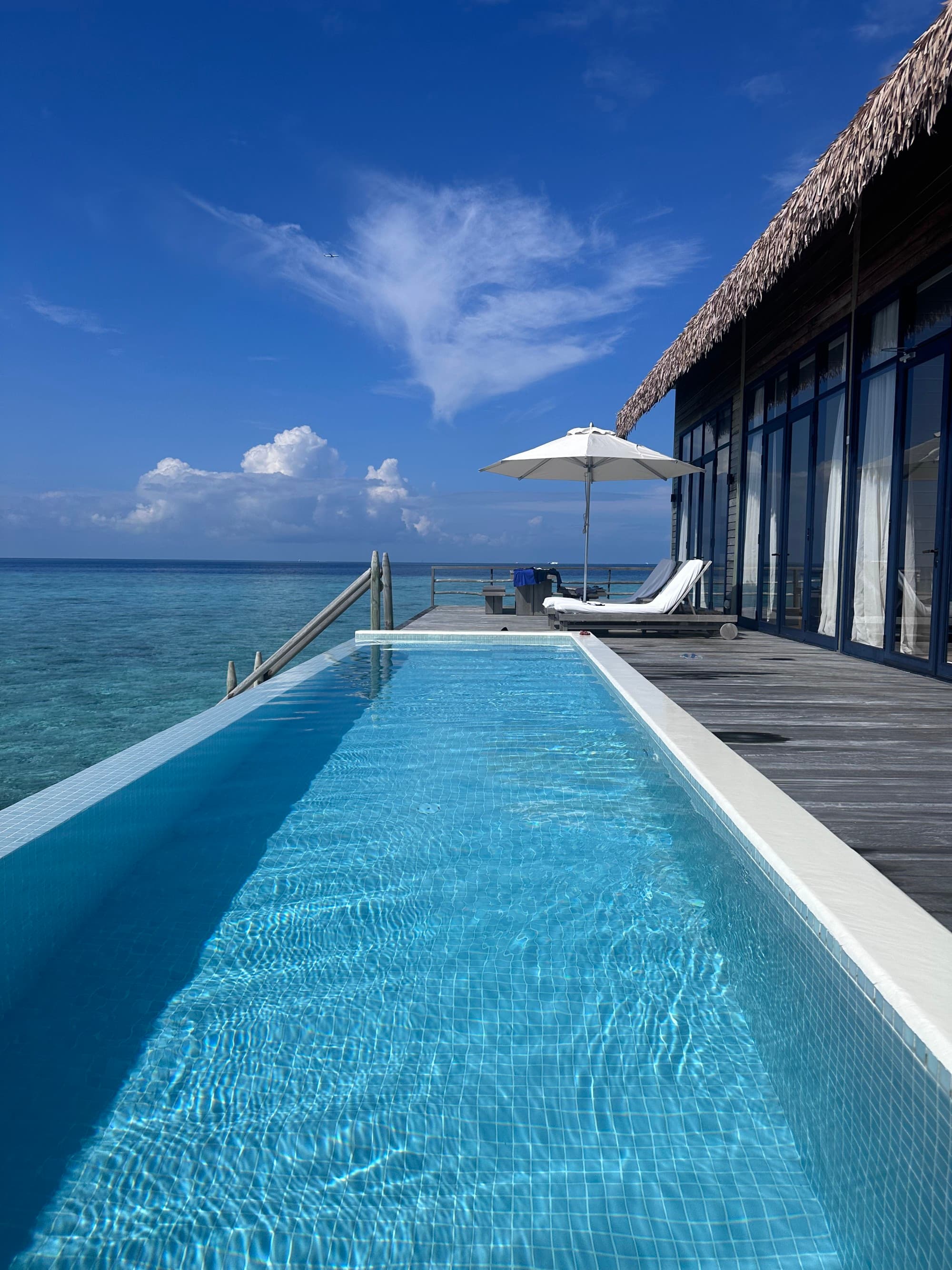 A long swimming pool next to a deck with an umbrella and lounge chair in the distance next to the sea.