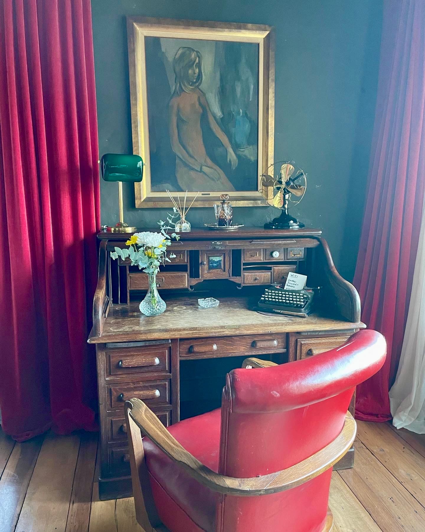 An old wooden desk with a red leather chair in front of it. There is a gold framed painting and red curtains on the wall, as well as home accessories on top of the desk.
