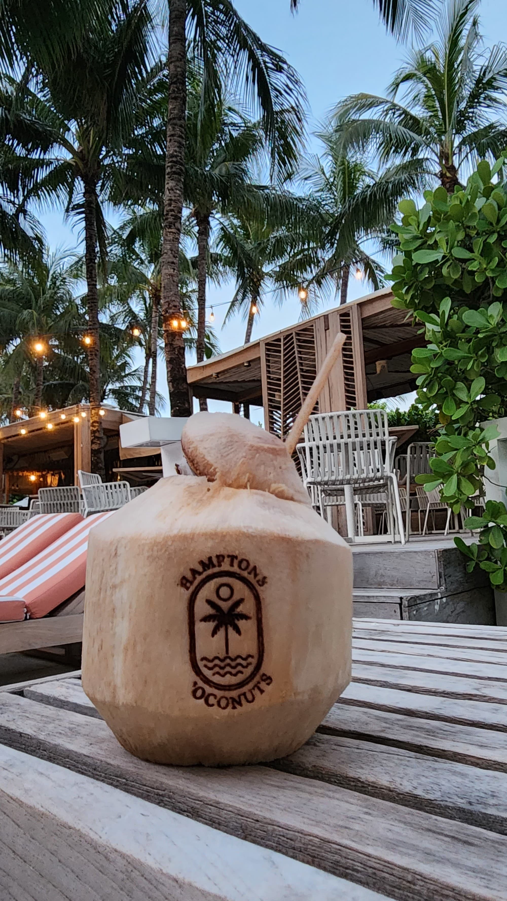 A coconut with an imprinted logo and straw on a table by the pool with palm trees