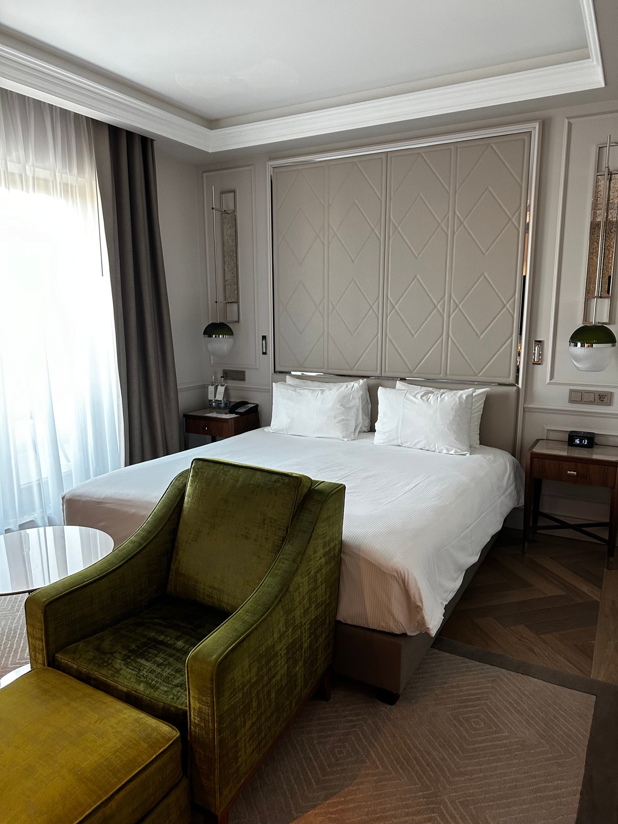 A hotel suite with a white bed, two night stands, elaborate sconces, a green chair, a carpet and curtains on the window on the left.